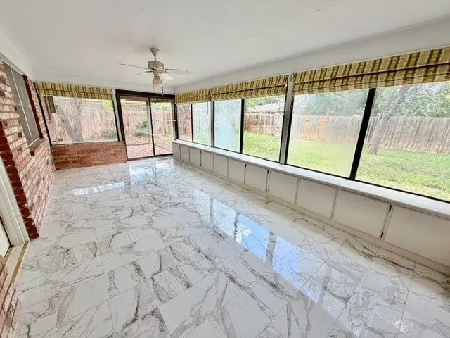 Sunroom featuring large-format marble-look tile flooring, extensive fenestration with horizontal blinds, built-in lower cabinetry, a ceiling fan, and exposed brick accents