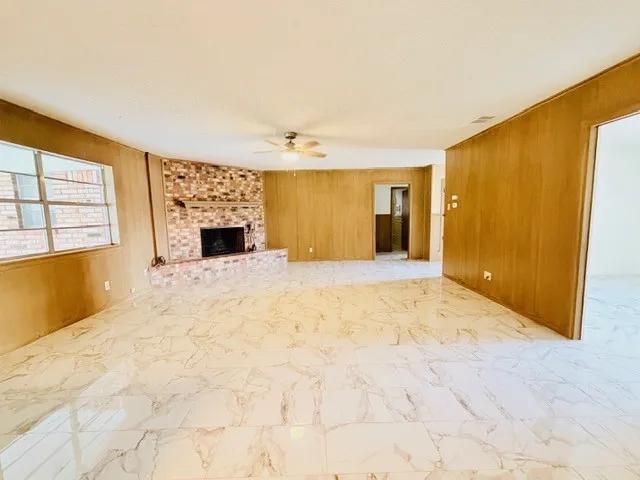 Spacious interior featuring marble-look tile flooring, a corner brick fireplace with hearth, wood paneling, a ceiling fan, and multiple window openings