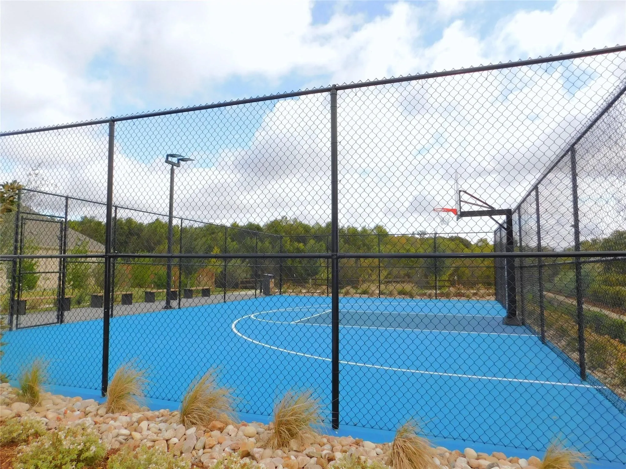 Fenced sport court featuring a blue playing surface with white line markings, a basketball hoop, and an overhead light fixture