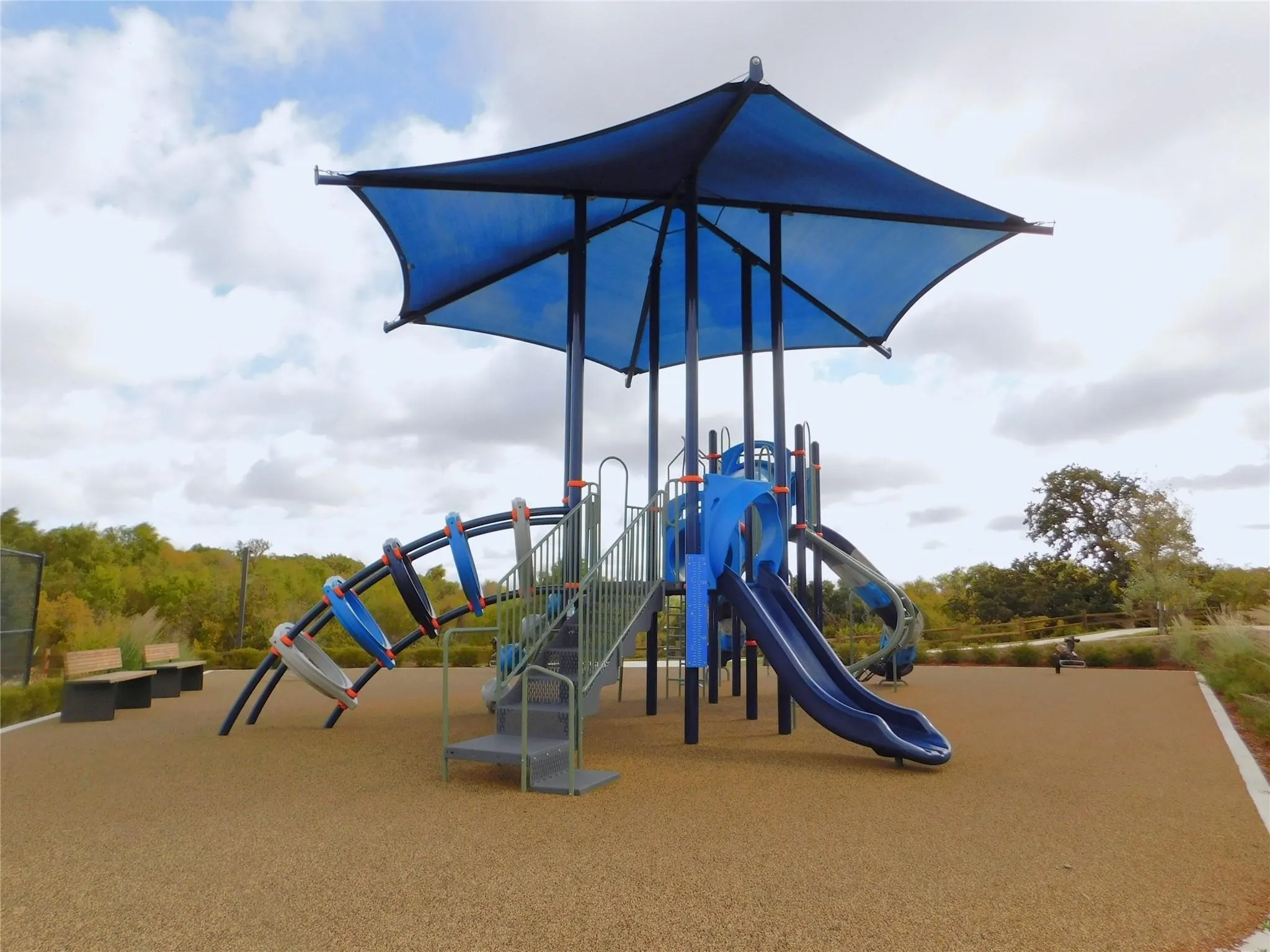Recreational playground area featuring a blue shade canopy, multiple slides, climbing structures, and a soft rubberized surface