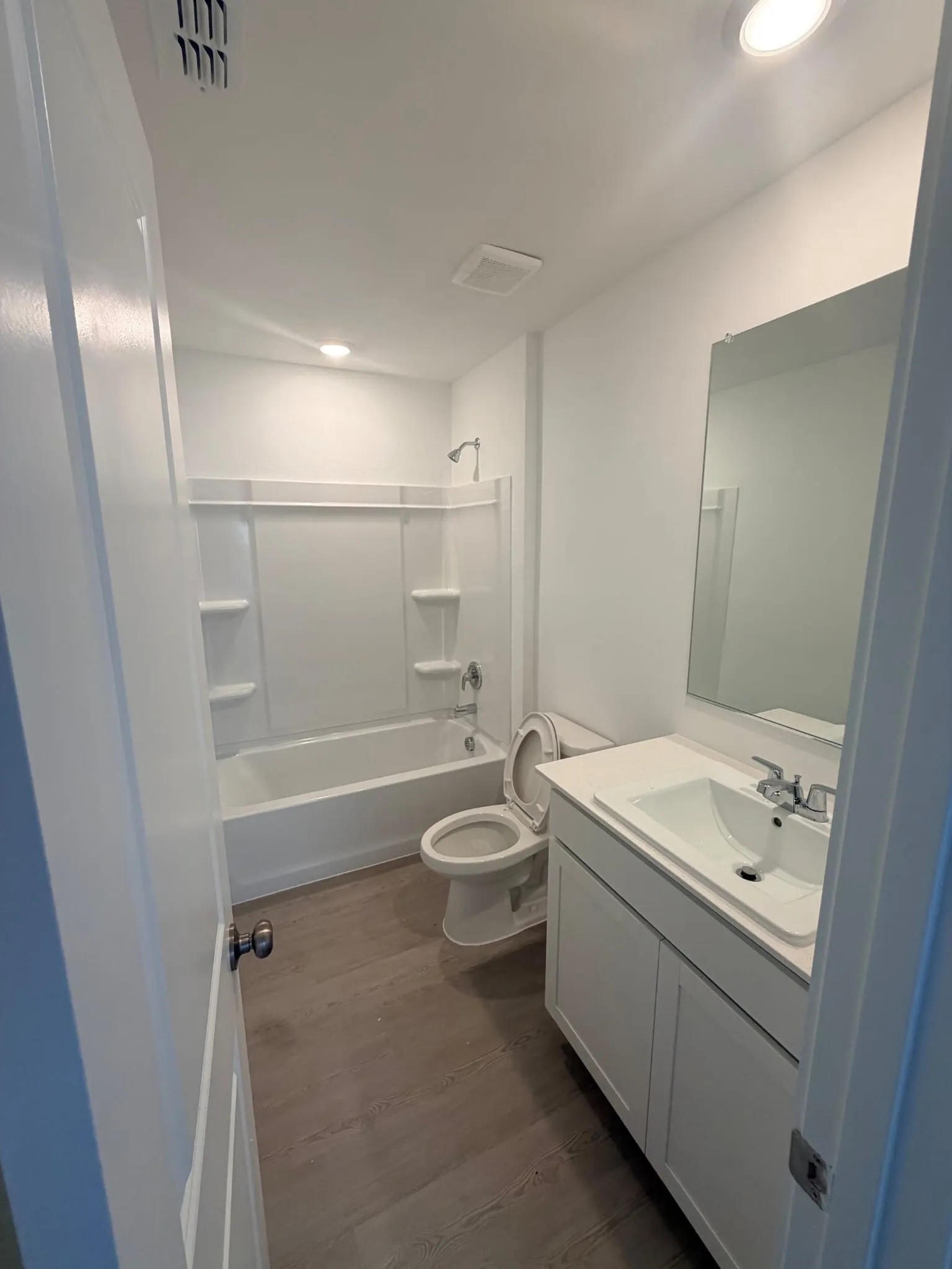 Bathroom featuring a white bathtub and shower combination with an integrated shelving unit, a white vanity with a single basin sink, a wall-mounted mirror, and wood-finish flooring