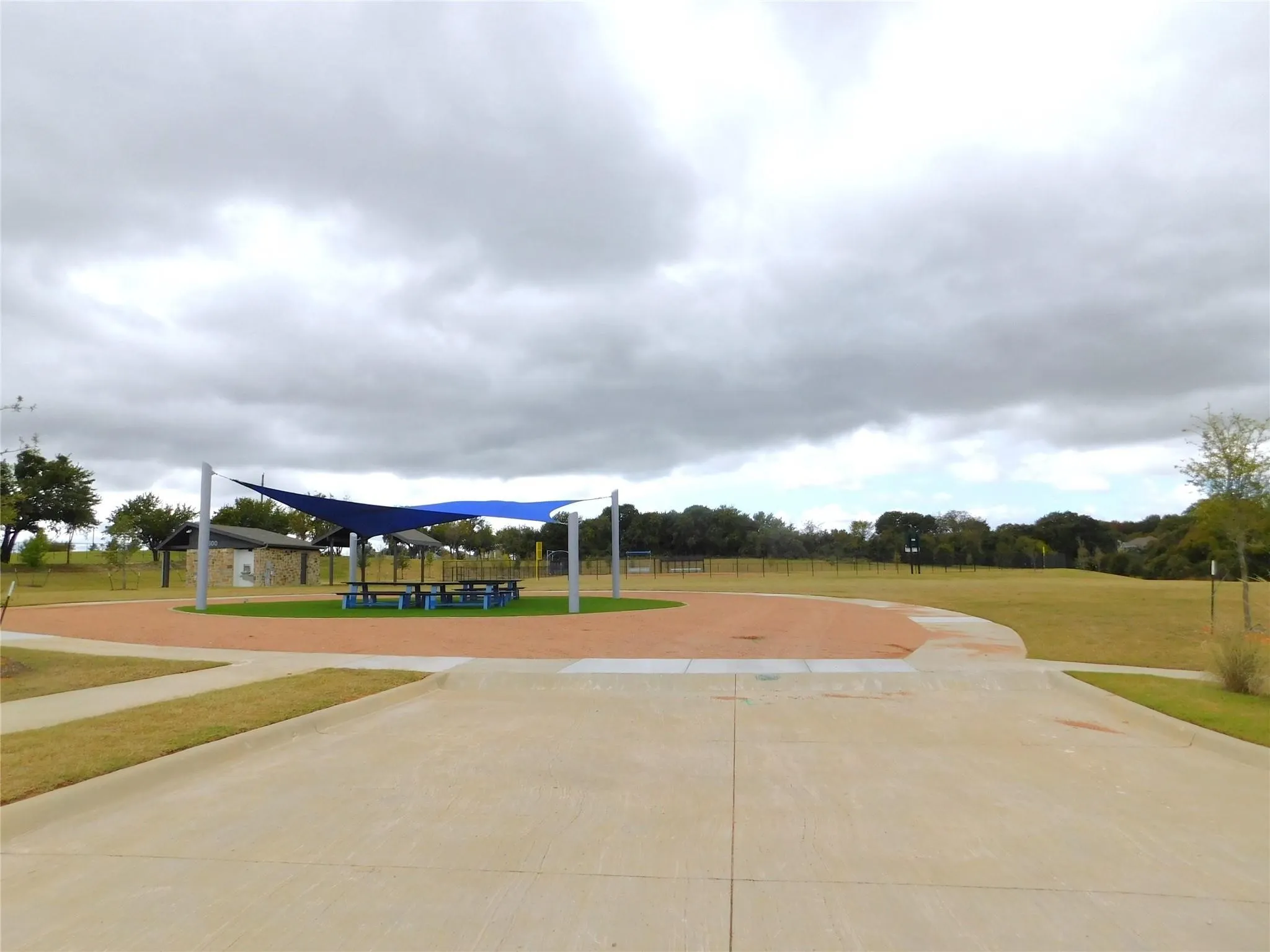 Community recreation area featuring shade structures, picnic tables, expansive concrete pathways, and a large open grassy field