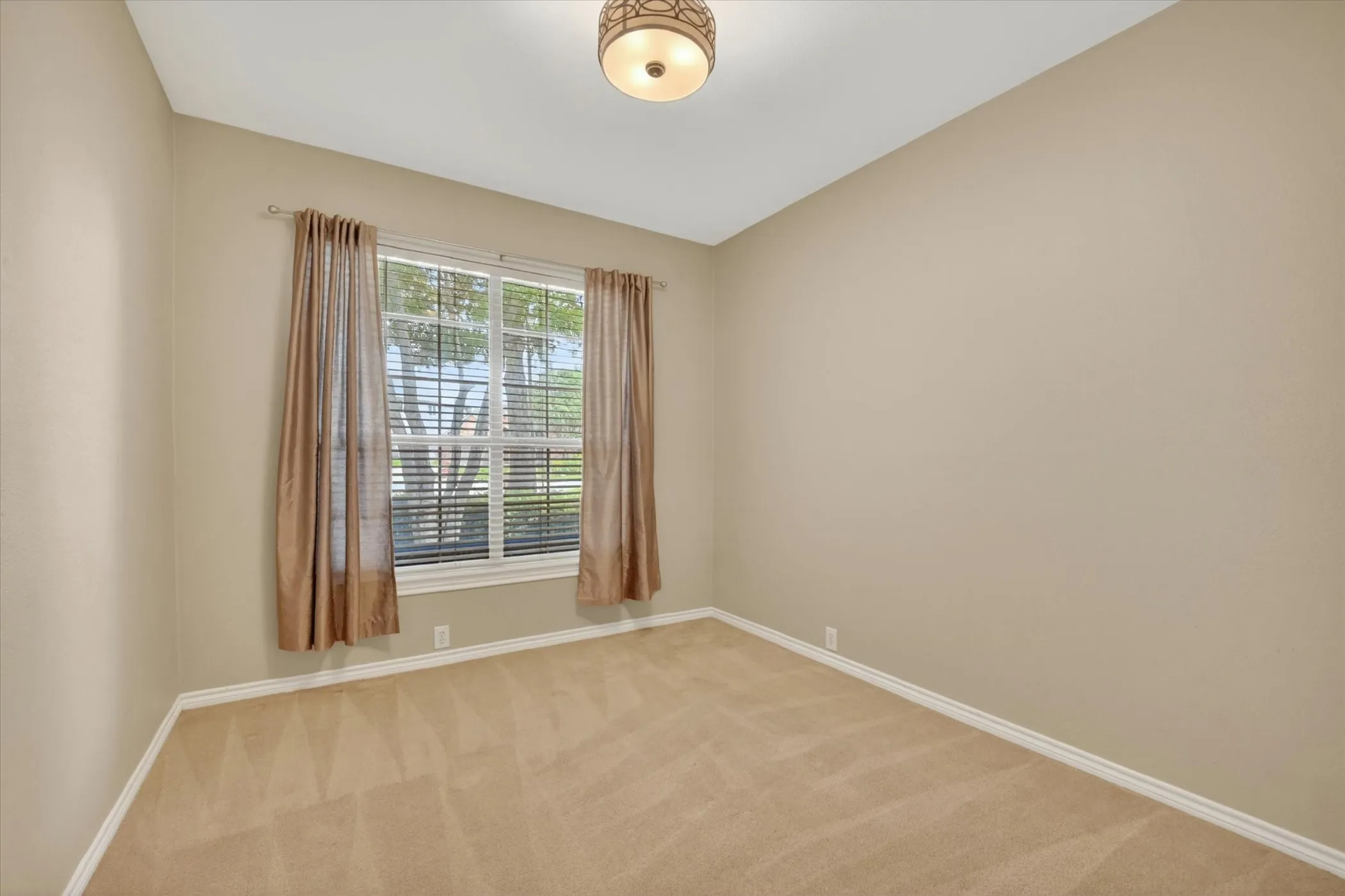 Neutral-toned room featuring a large window with blinds and drapes, beige carpeting, light-colored walls, and a decorative ceiling light fixture