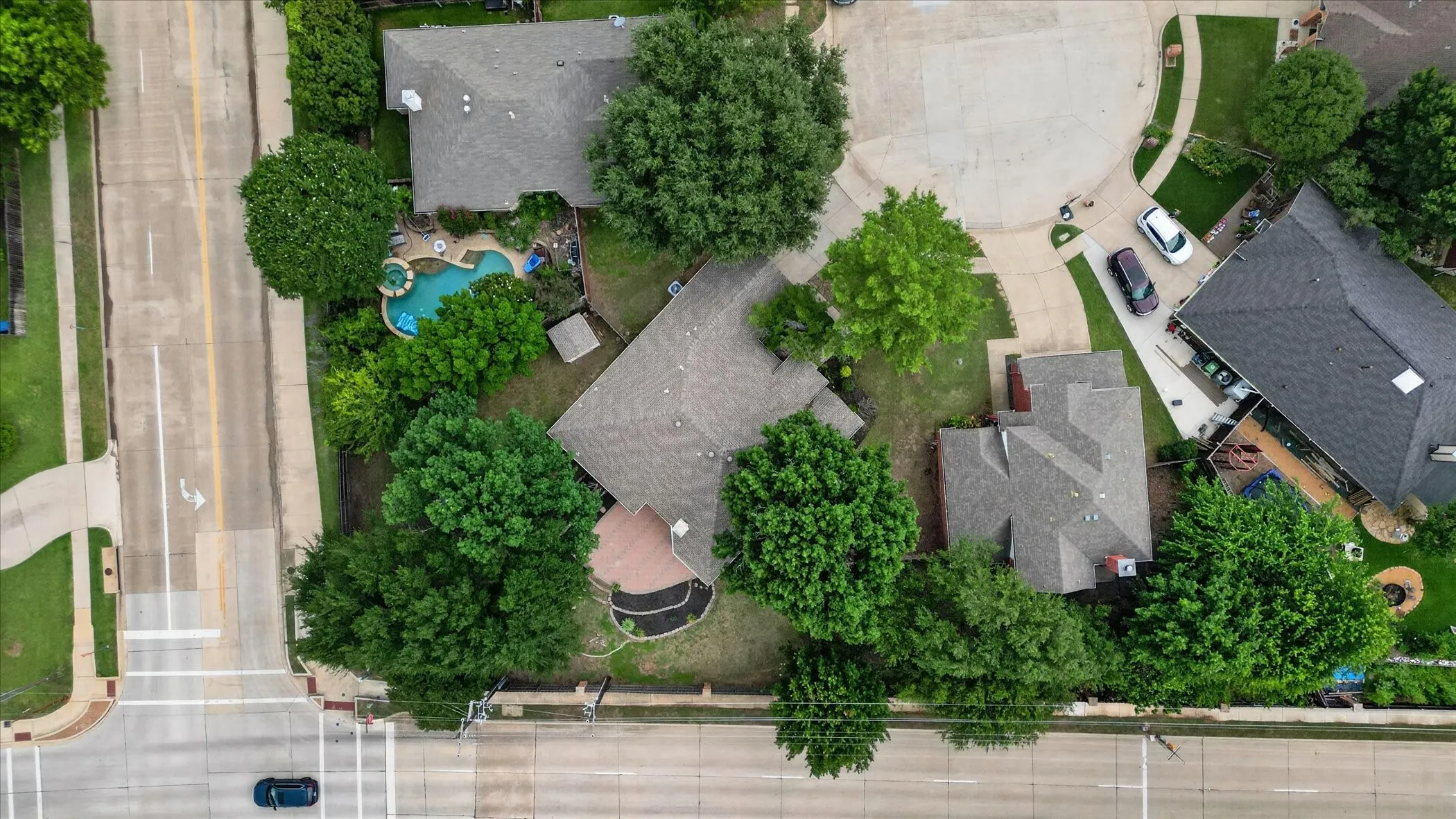 Aerial perspective showcasing a property featuring a curvilinear swimming pool, mature perimeter trees, and a paved patio area