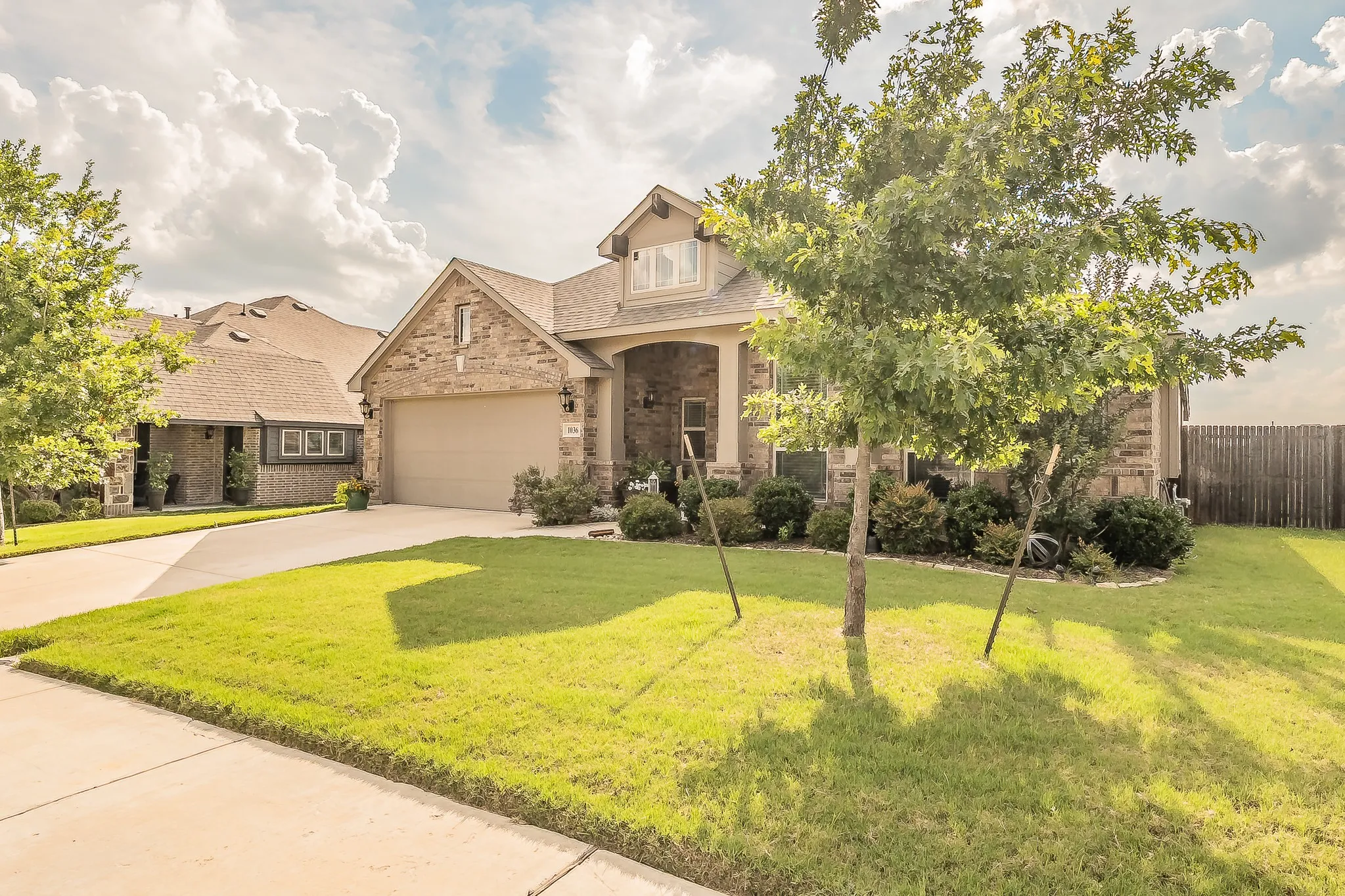 Stone and stucco exterior featuring an attached two-car garage, covered porch, well-maintained lawn, established landscaping, and a privacy fence