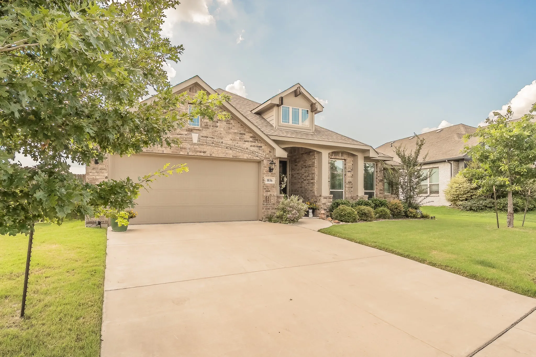 Brick and stone facade residence featuring a covered entry, a prominent dormer, an attached garage, a concrete driveway, and manicured landscaping