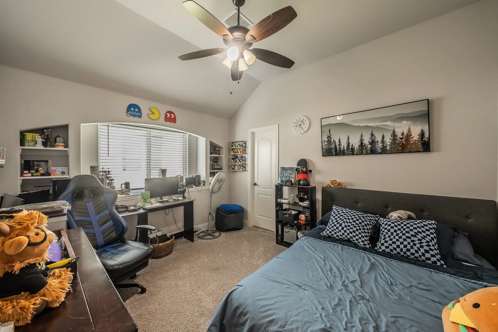 Carpeted room featuring a vaulted ceiling with a ceiling fan, a window with horizontal blinds, built-in shelving, and a solid core door