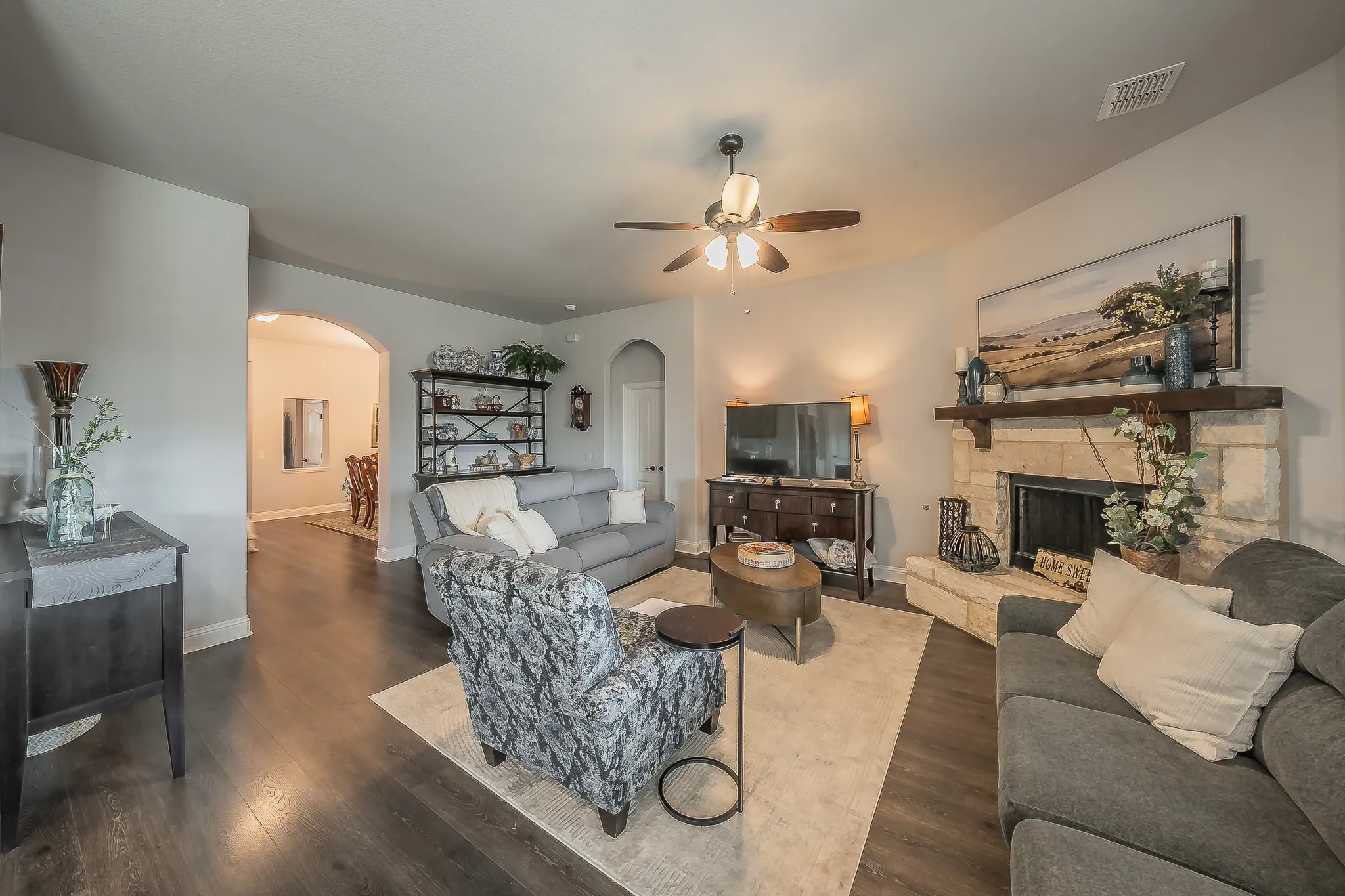 Spacious living area featuring wood-finish flooring, a stone fireplace with a substantial wood mantel, a ceiling fan, and arched doorways