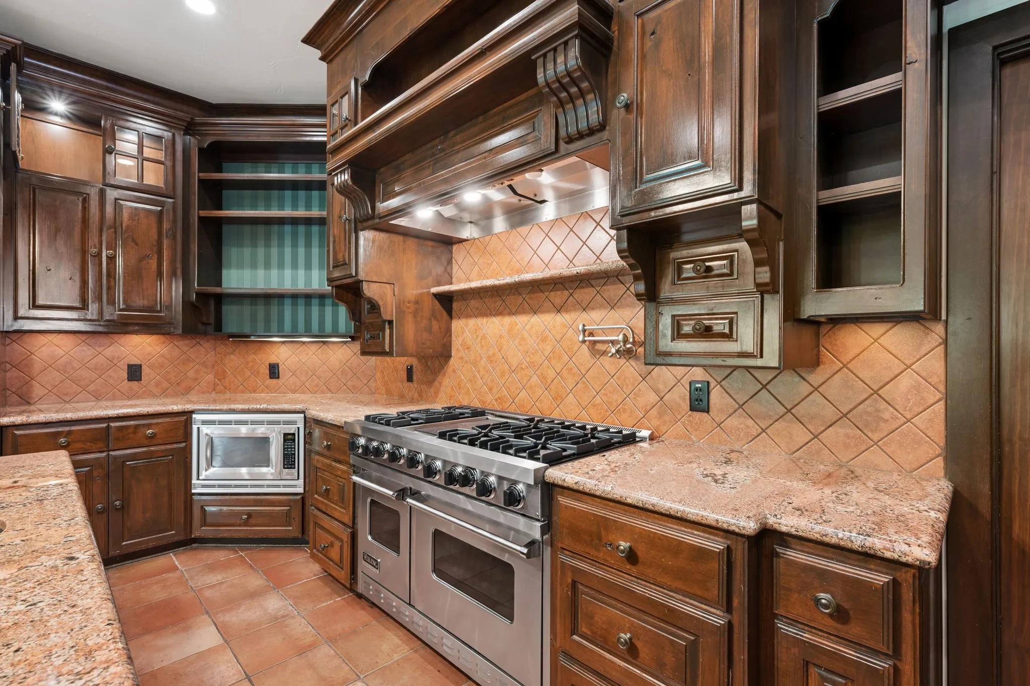 Gourmet kitchen featuring custom wood cabinetry, stainless steel range with a built-in microwave, granite countertops, and terra cotta tile flooring