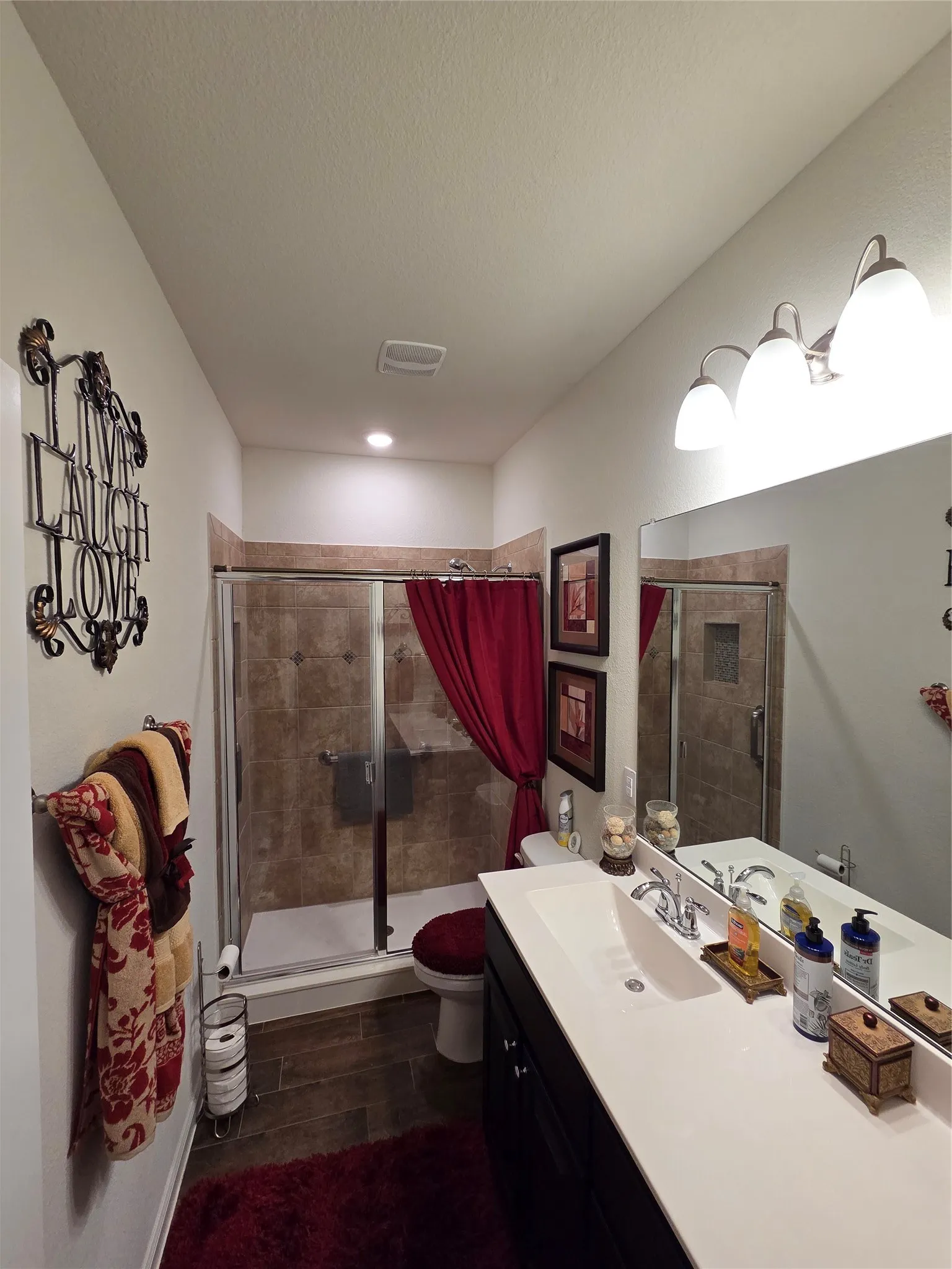Bathroom featuring a single vanity with a white countertop and integrated sink, a large wall-mounted mirror, wood-finish flooring, and a glass-enclosed shower with tiled walls