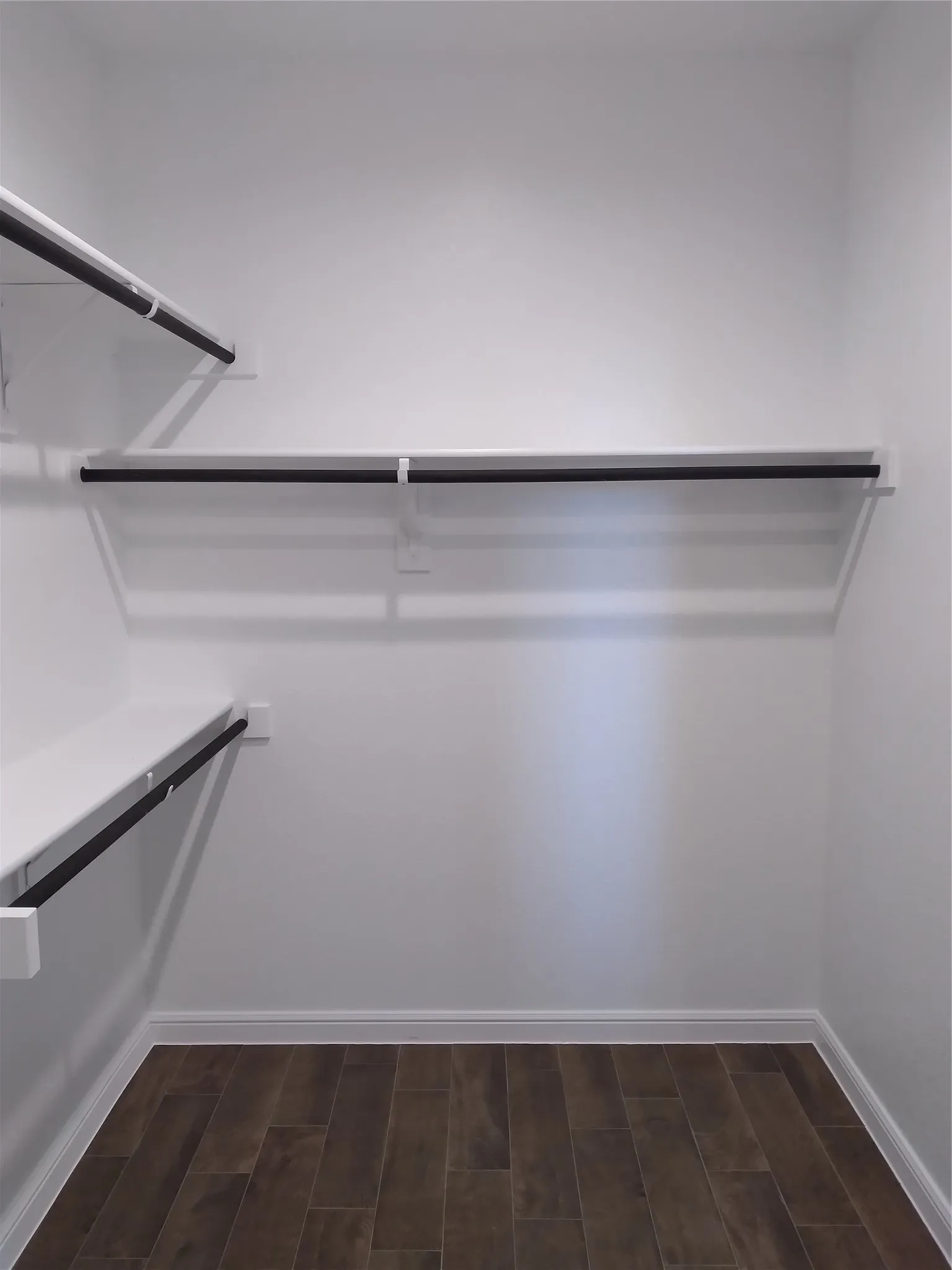 Walk-in closet featuring white walls, wood-finish flooring, white baseboards, and built-in white shelving with black hanging rods