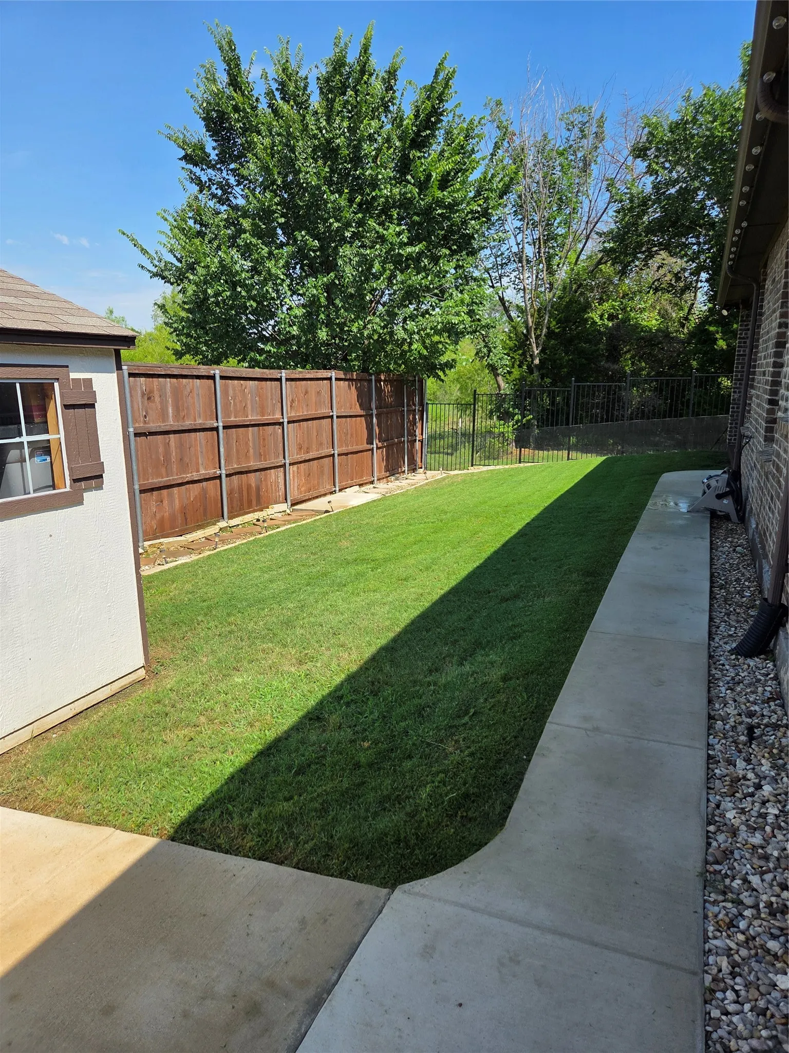 Expansive grassy area featuring a wood privacy fence, a black metal gate, concrete pathways, and a brick exterior wall