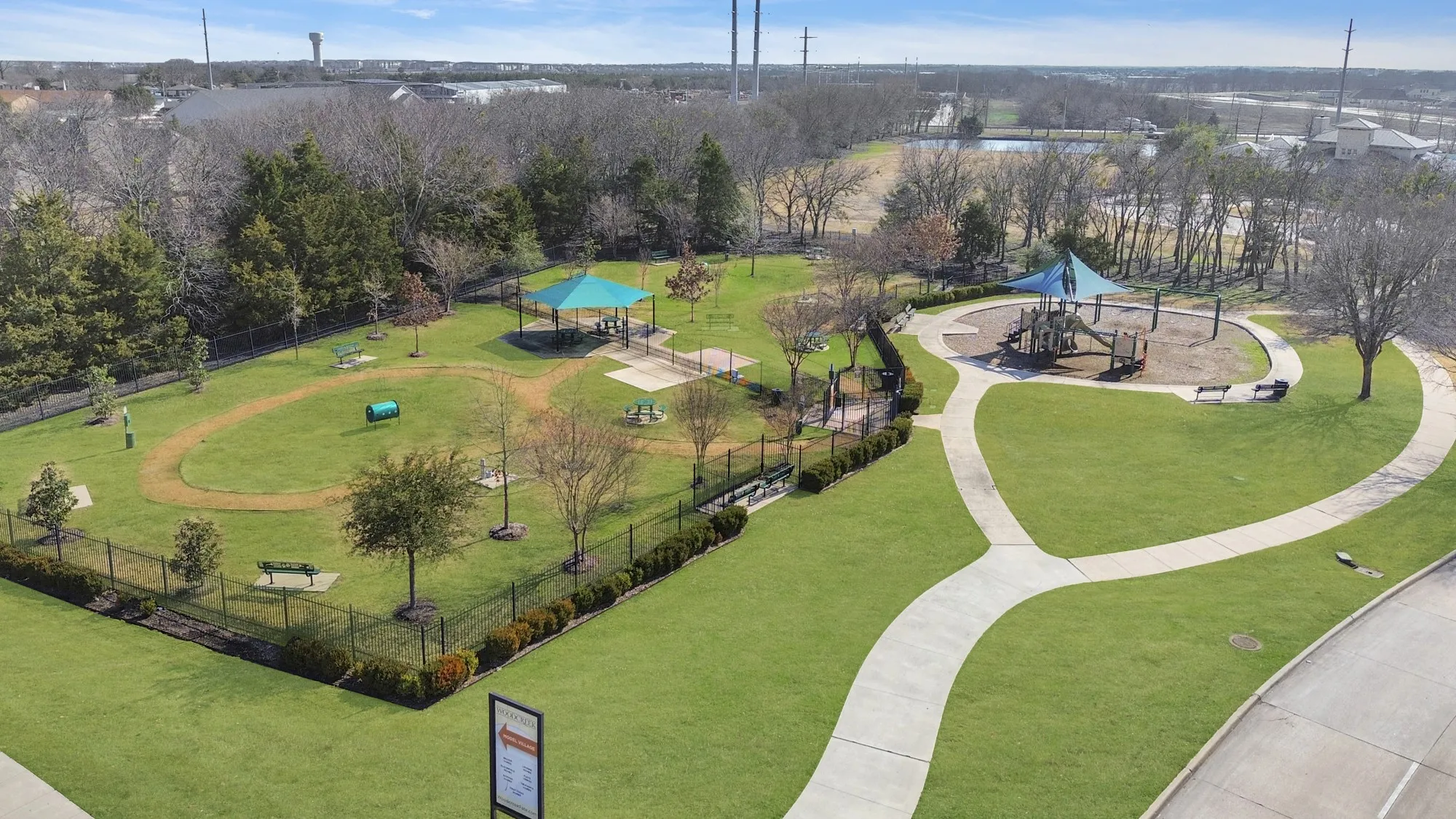 Expansive green space featuring a shaded playground, a fenced pet park with an agility tunnel, a covered picnic area, and paved walking paths