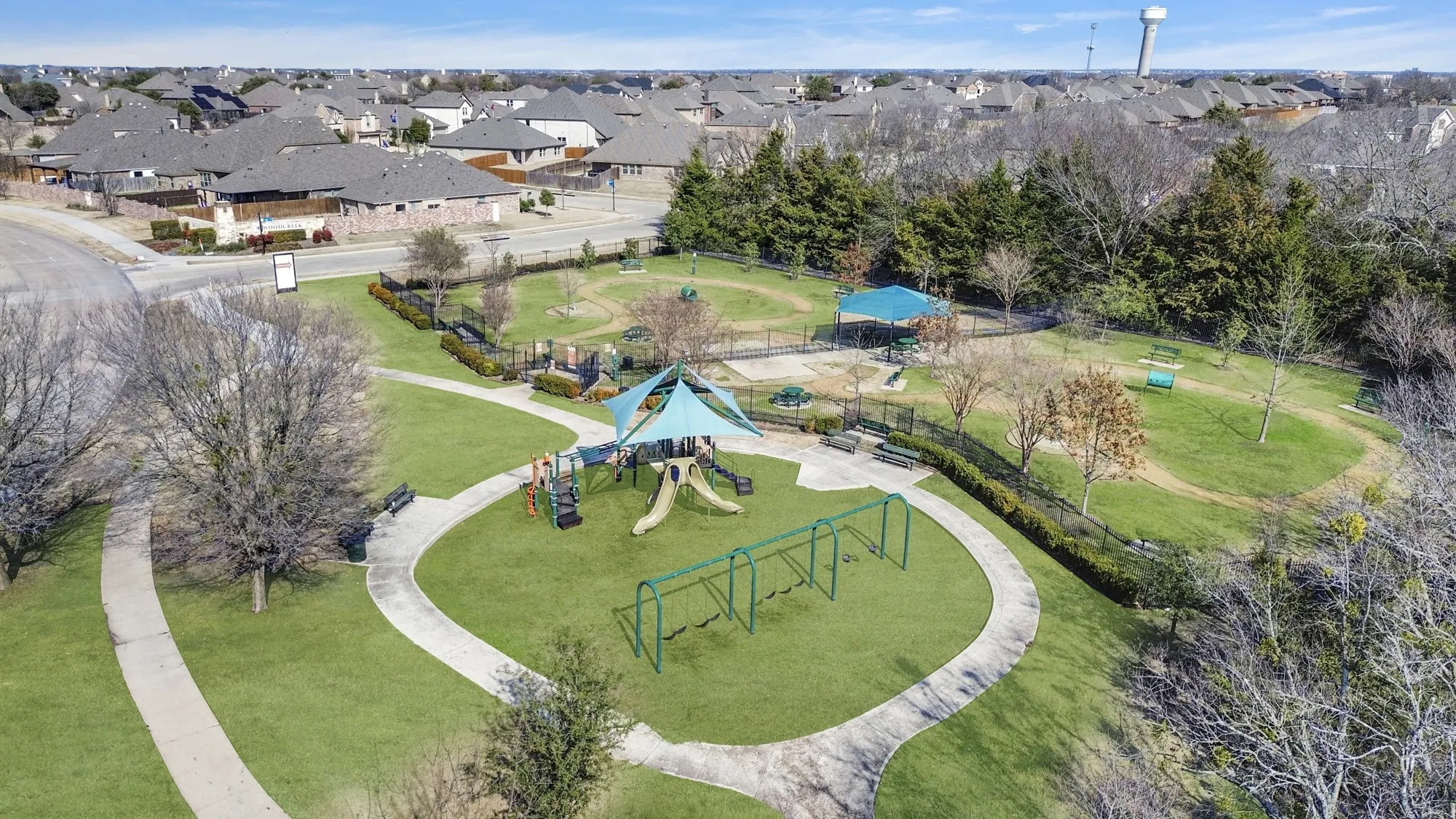 Expansive green space featuring a playground with slides and swings, shaded seating areas, winding concrete pathways, and surrounding residential properties
