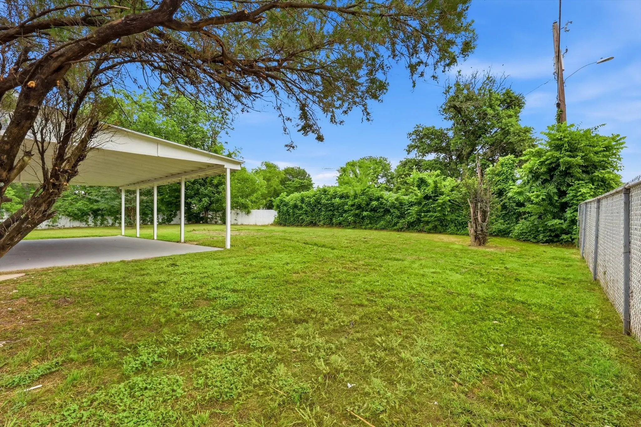 Expansive exterior featuring a carport with a concrete pad, a mature shade tree, a chain-link fence, and a large grassy​​‌​​​​‌​​‌‌​‌‌​​​‌‌​‌​‌​‌​​​‌​​ area
