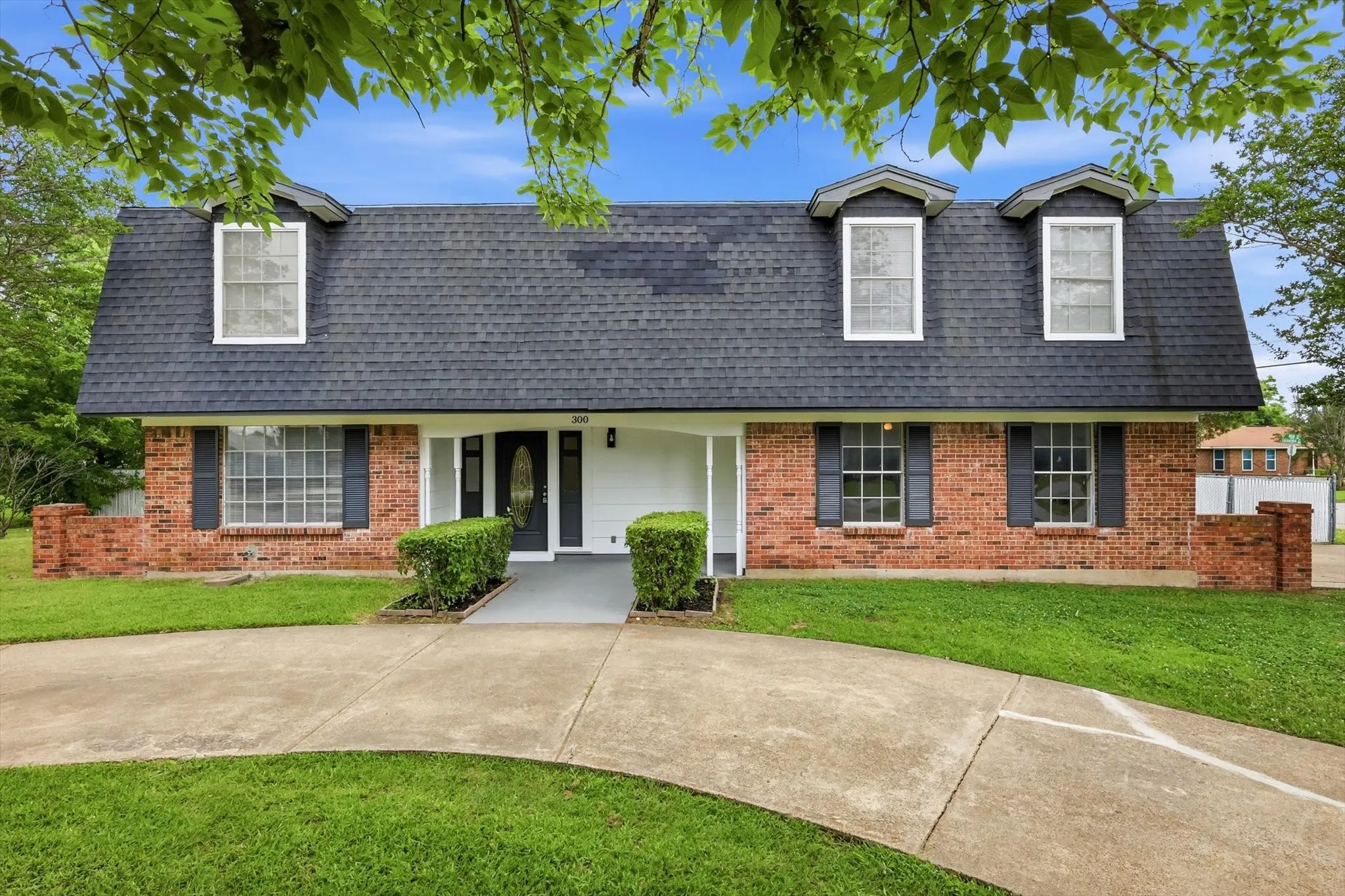 Brick facade residence featuring a dark shingle roof, three dormer windows, decorative shutters, and an arched front entry