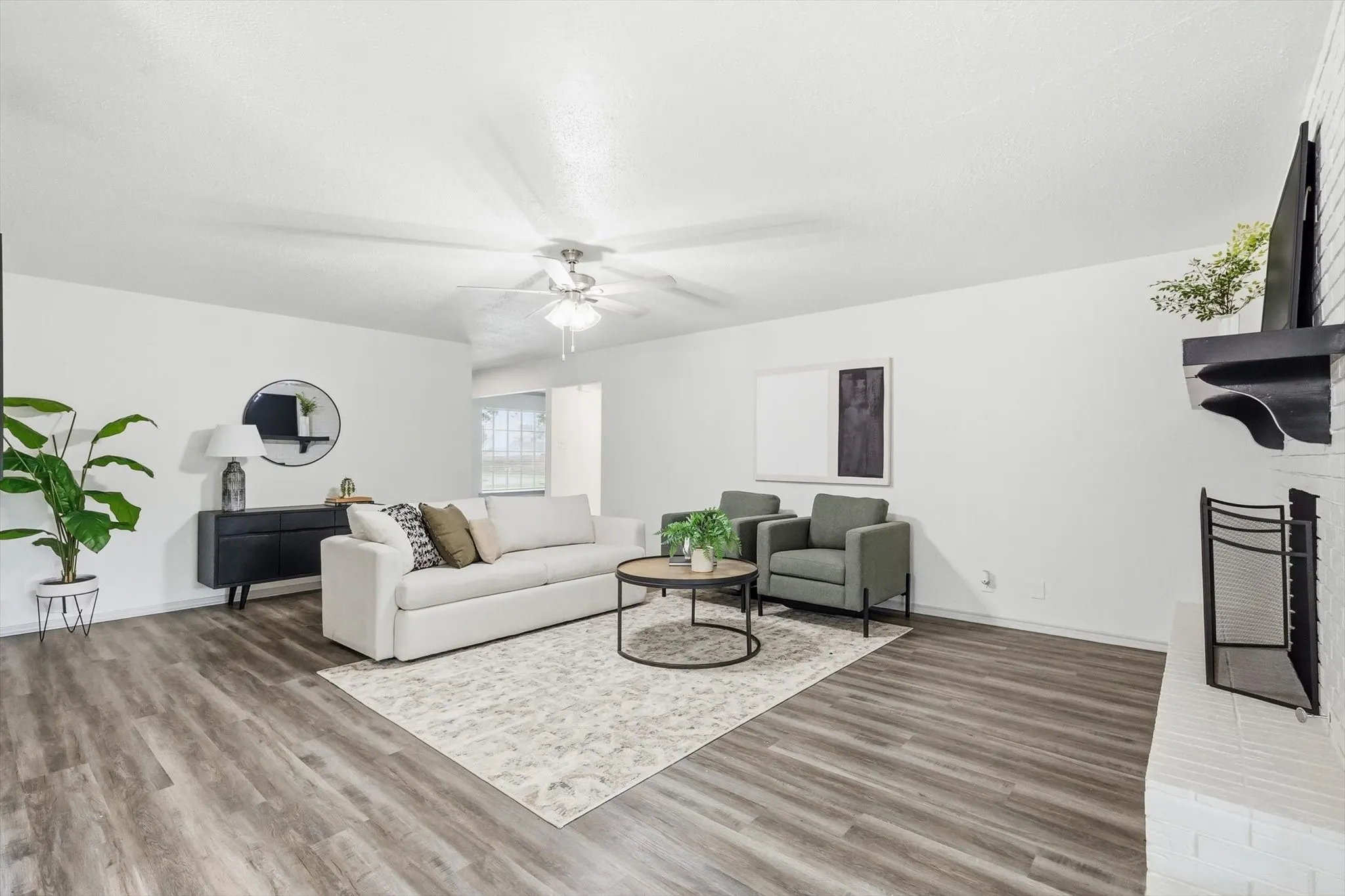 Spacious living area featuring wood-finish flooring, a white brick fireplace with a black mantel, and a ceiling fan with integrated lighting