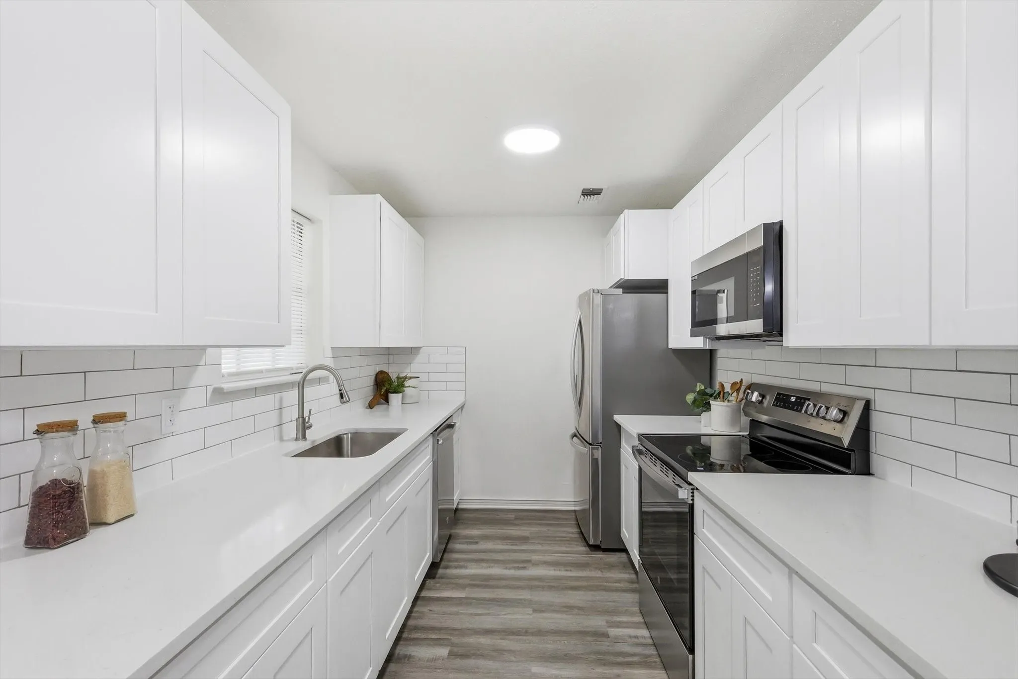 Galley kitchen featuring white shaker cabinetry, stainless steel appliances, white subway tile backsplash, light-toned countertops, and wood-finish flooring
