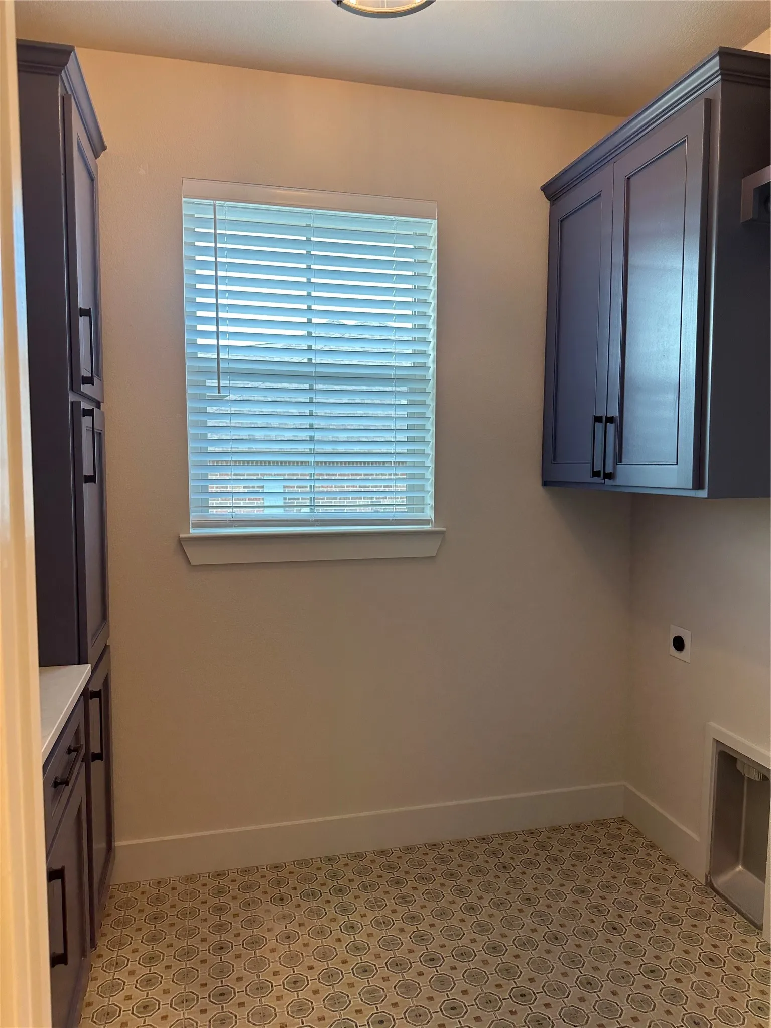 Utility space featuring patterned floor tile, built-in cabinetry with dark hardware, a window with blinds, and white baseboards