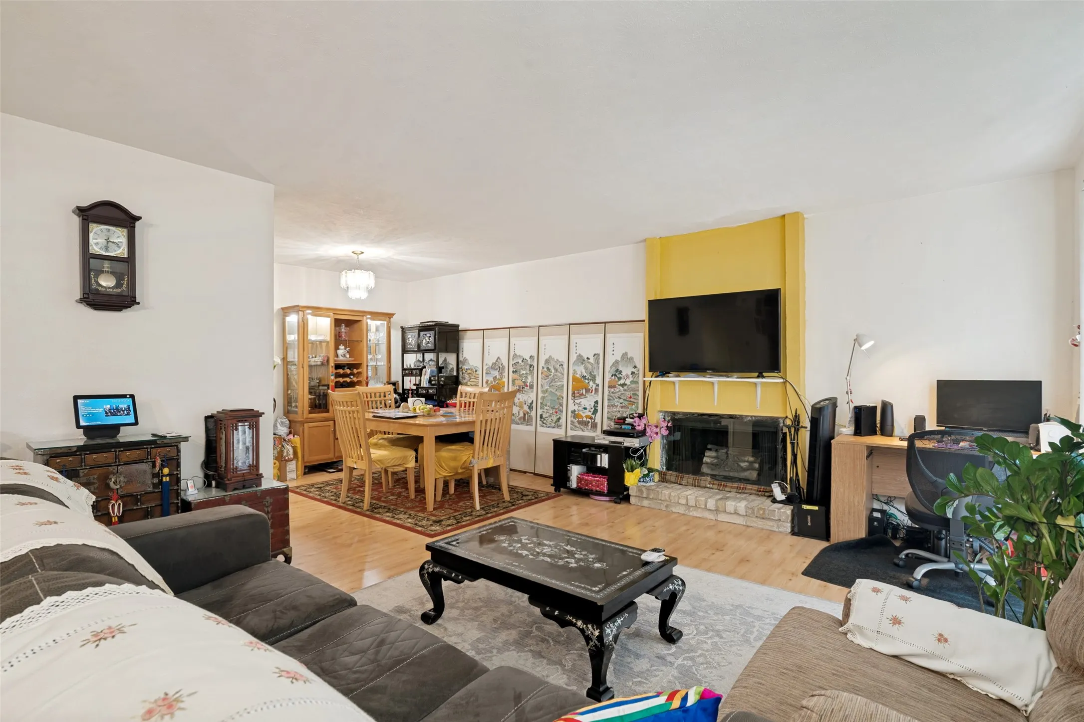 Open-concept living area featuring wood-finish flooring, a brick-surround fireplace with a yellow accent wall, and a distinct dining area with chandelier lighting
