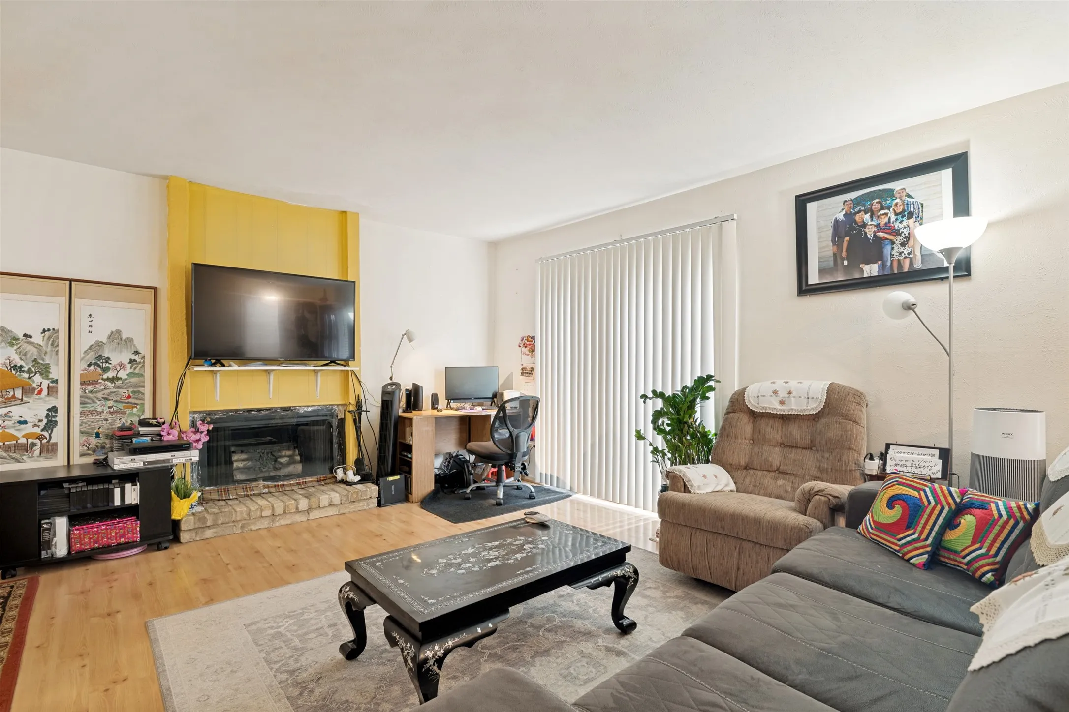 Living area featuring wood-finish flooring, a brick fireplace with a yellow accent wall, and vertical blinds