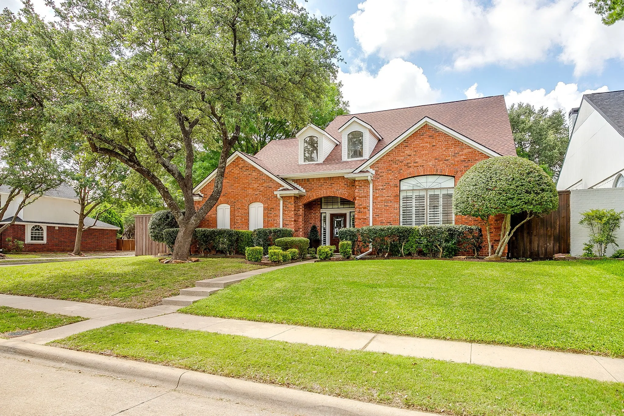 Brick facade residence featuring a front lawn, mature trees, landscaping, and an arched entry