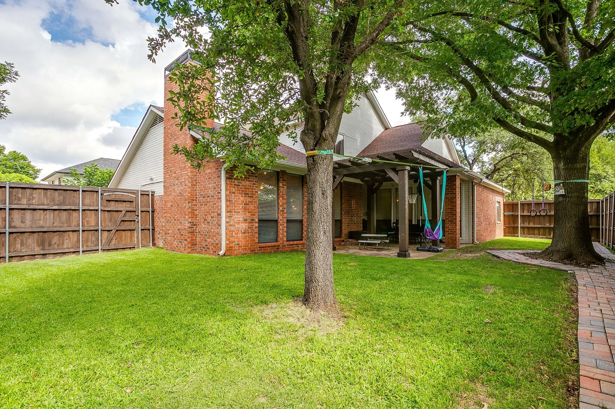Spacious backyard featuring a covered patio, extensive lawn, mature trees, brick paver walkway, and privacy fencing