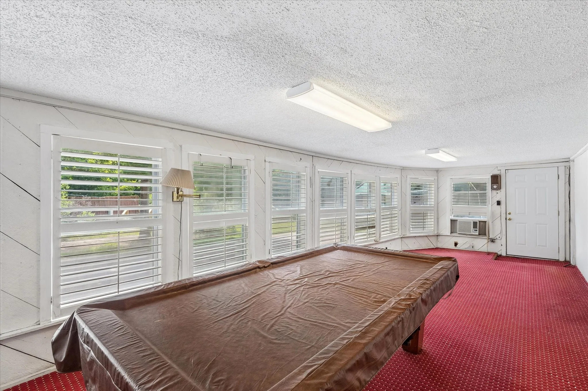 Sunroom featuring multiple windows with louvered shutters, red patterned carpeting, and white textured ceiling