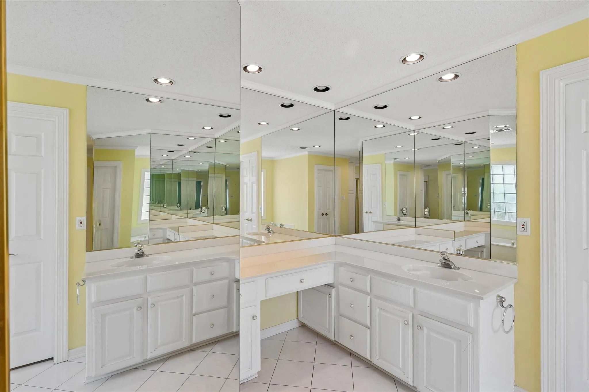 Bathroom featuring extensive vanity space with dual integrated sinks, white cabinetry, a built-in makeup area, comprehensive wall-to-wall mirrors, and recessed ceiling lighting