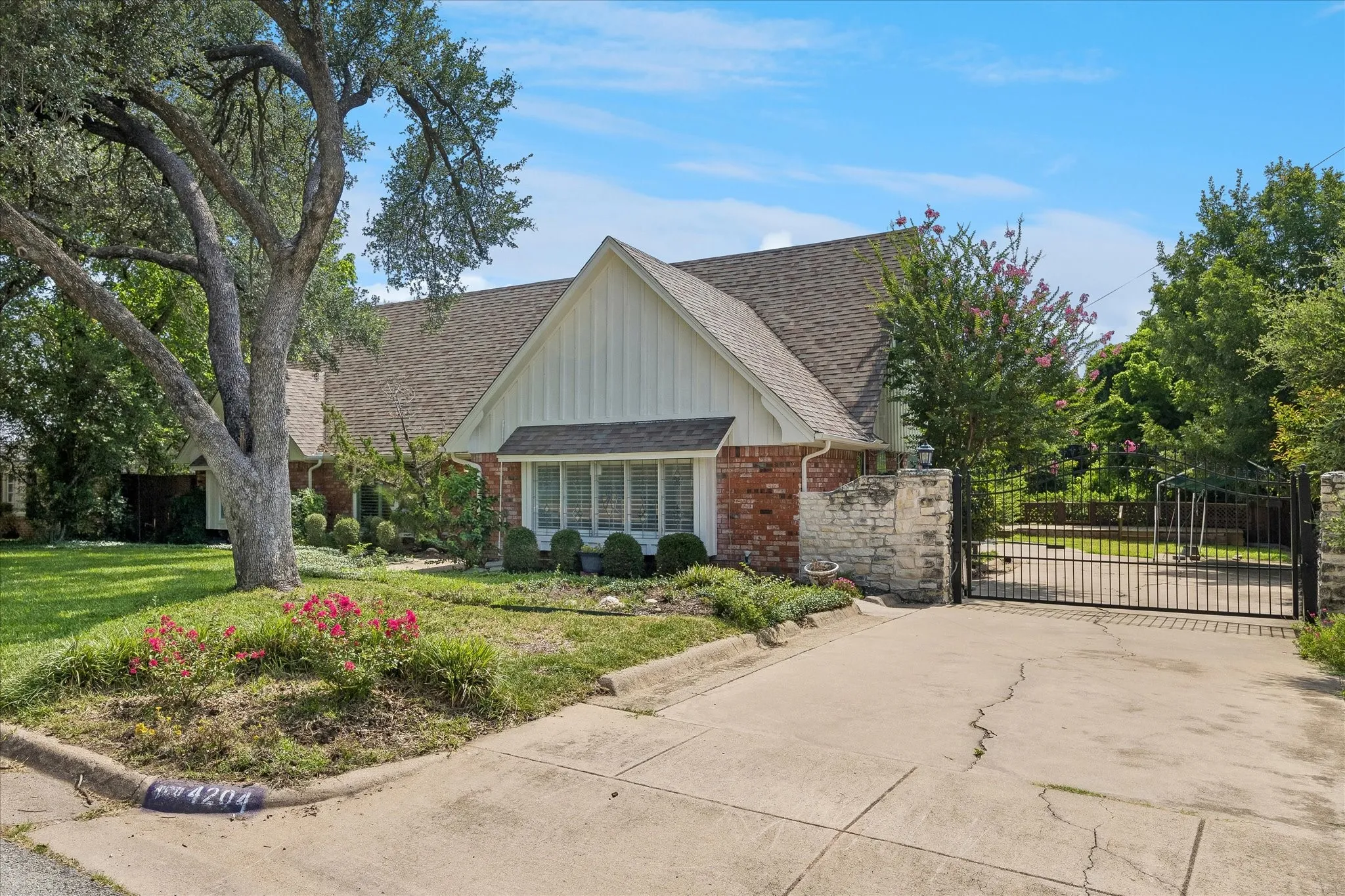 Brick and vertical siding exterior featuring a multi-gable roofline, a front lawn with mature trees, and a concrete driveway leading to a metal gate with stone pillars