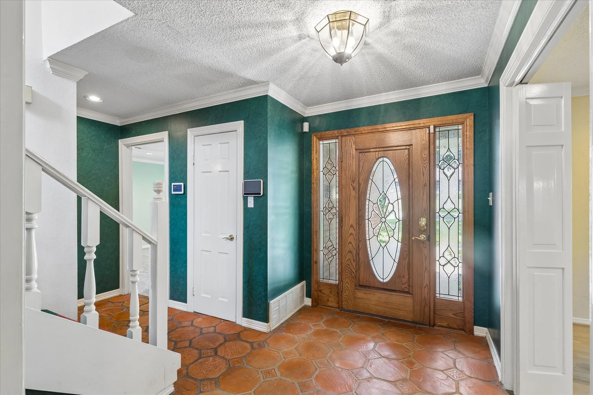 Foyer featuring a wood-panel entry door with oval and rectangular decorative glass inserts, terracotta-toned hexagonal floor tiles, and a white baluster staircase