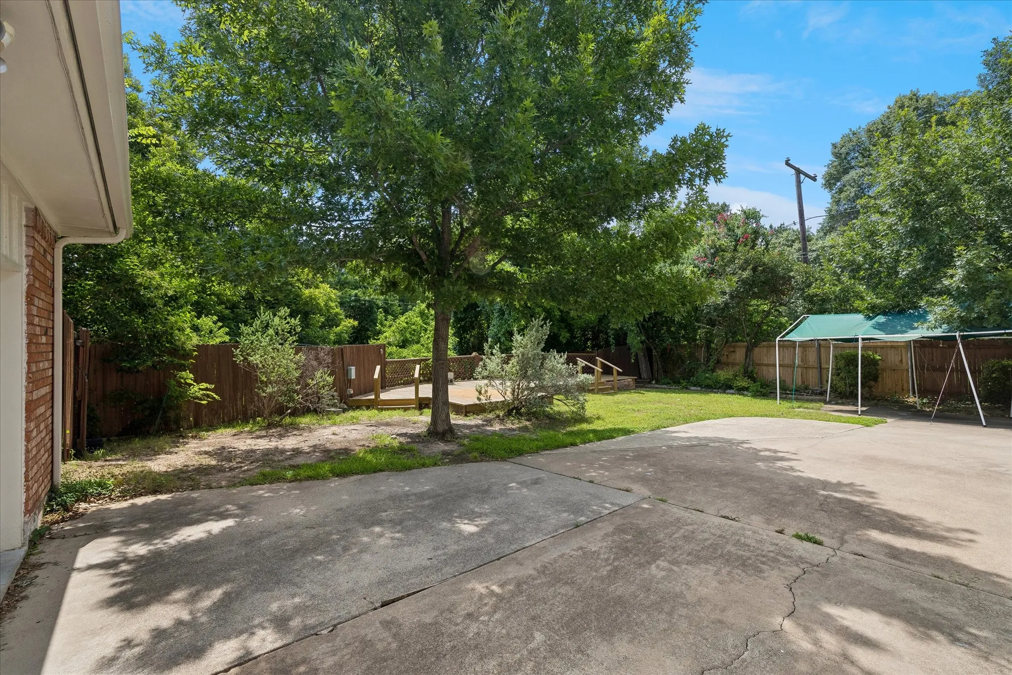 Expansive concrete patio with a wooden deck, mature trees, a wooden privacy fence, and an artificial turf covered awning