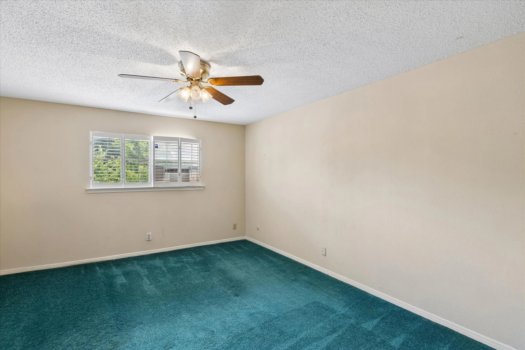 Spacious room featuring neutral wall paint, textured ceiling, a ceiling fan with light fixture, and a window with white plantation shutters