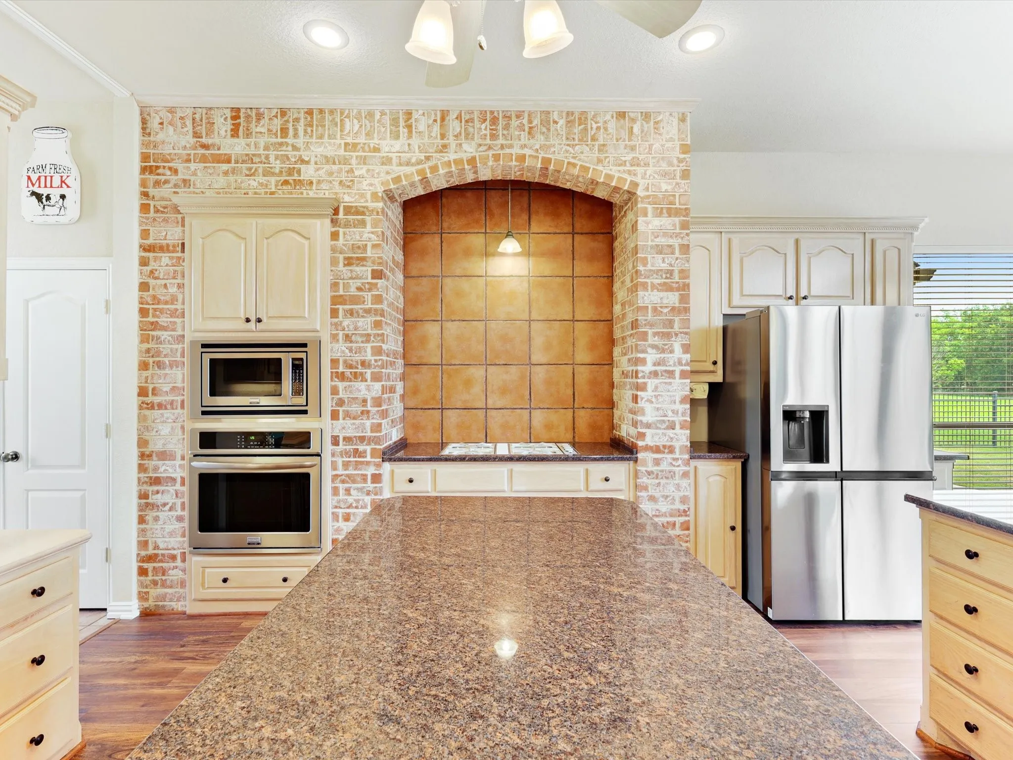 Spacious kitchen featuring a large island with a patterned countertop, a brick-faced wall with an arched alcove, built-in stainless steel appliances including an oven and microwave, light-toned cabinetry, and wood-finish flooring