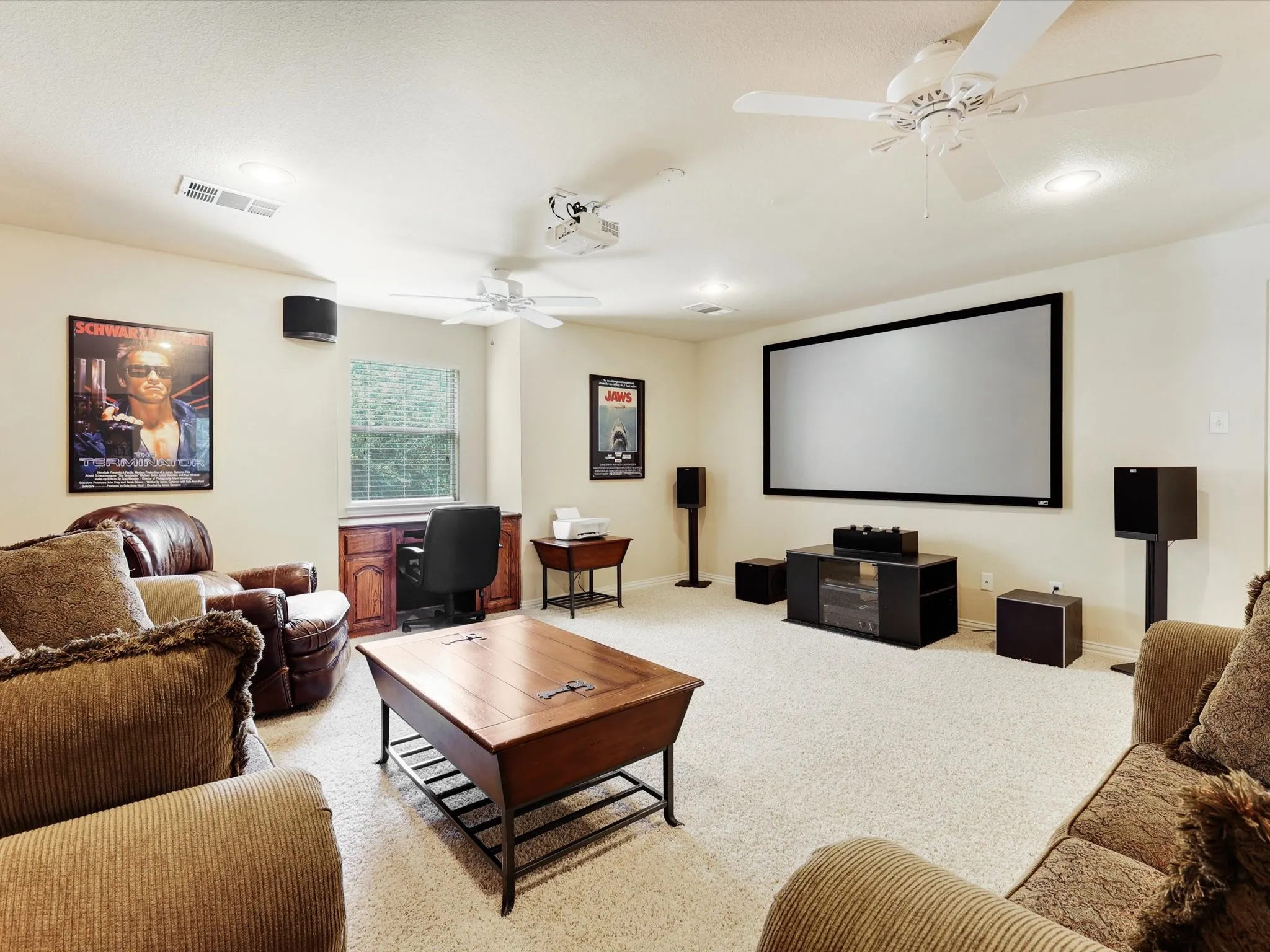 Spacious media room featuring a large projection screen, built-in desk with wood cabinetry, recessed lighting, and ceiling fans