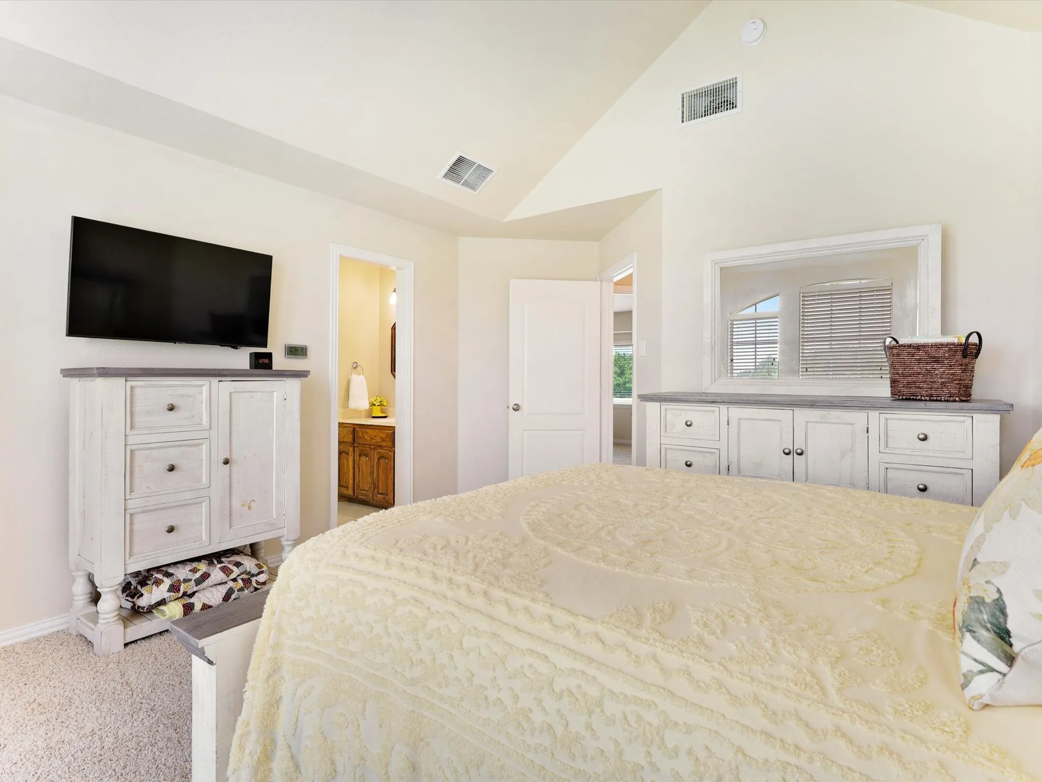 Vaulted ceiling bedroom featuring plush carpeting, white trim, and a doorway opening to a bathroom with a wood-finish vanity