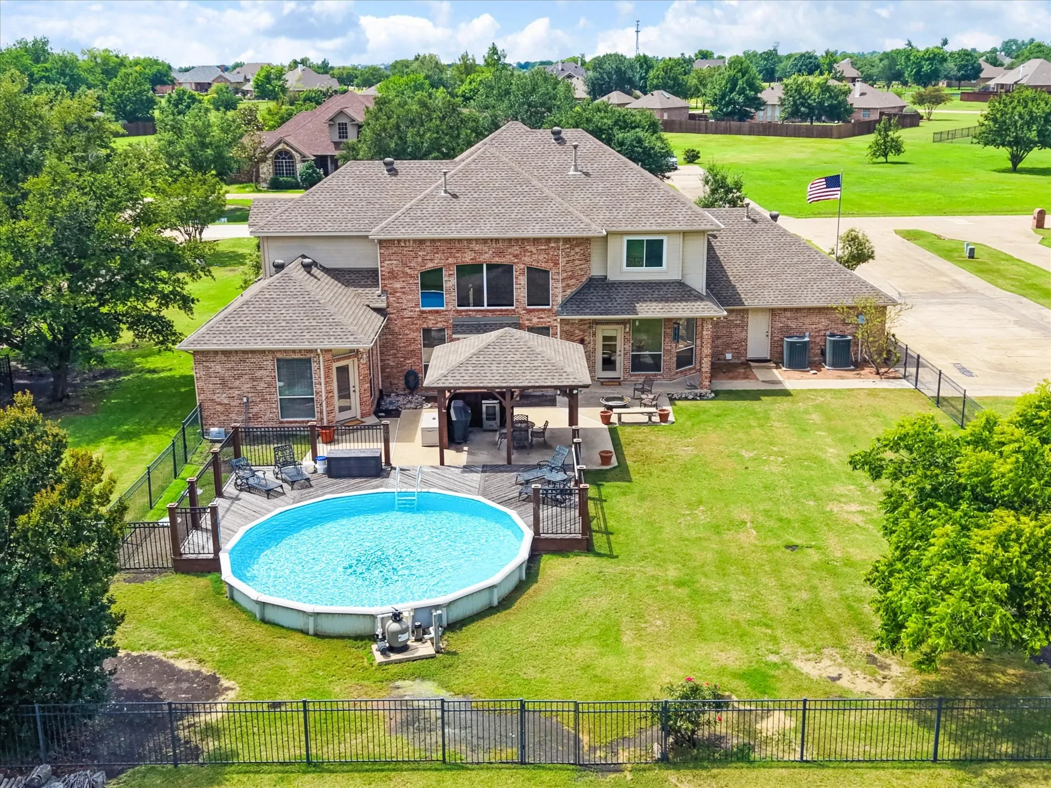 Expansive rear exterior featuring a multi-level brick facade with multiple windows, a large above-ground pool with surrounding wood decking, a covered patio area, and a substantial fenced yard
