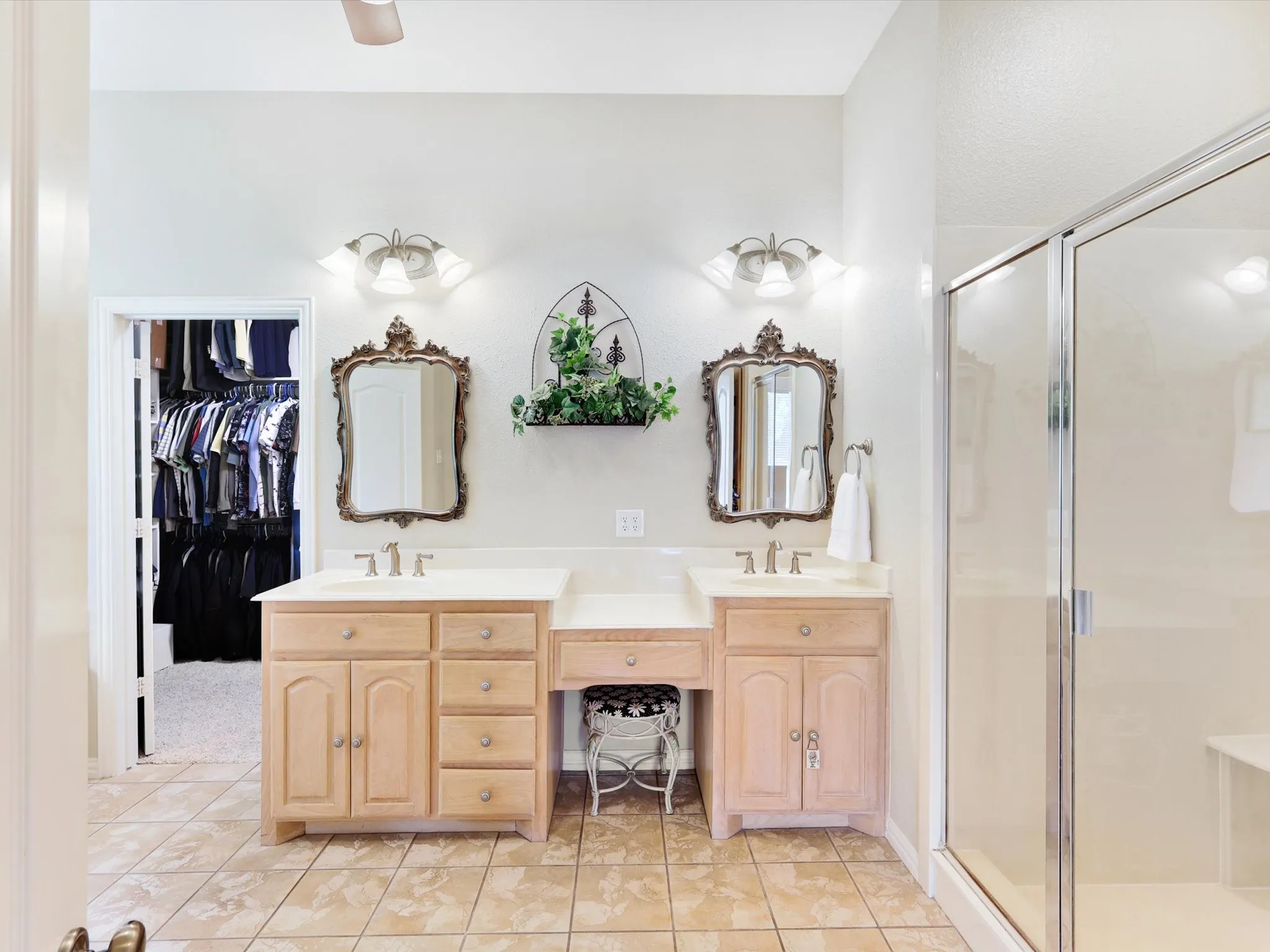 Bathroom featuring dual vanities with light wood cabinetry and countertops, a glass-enclosed shower with an integrated bench, and a walk-in closet with built-in shelving