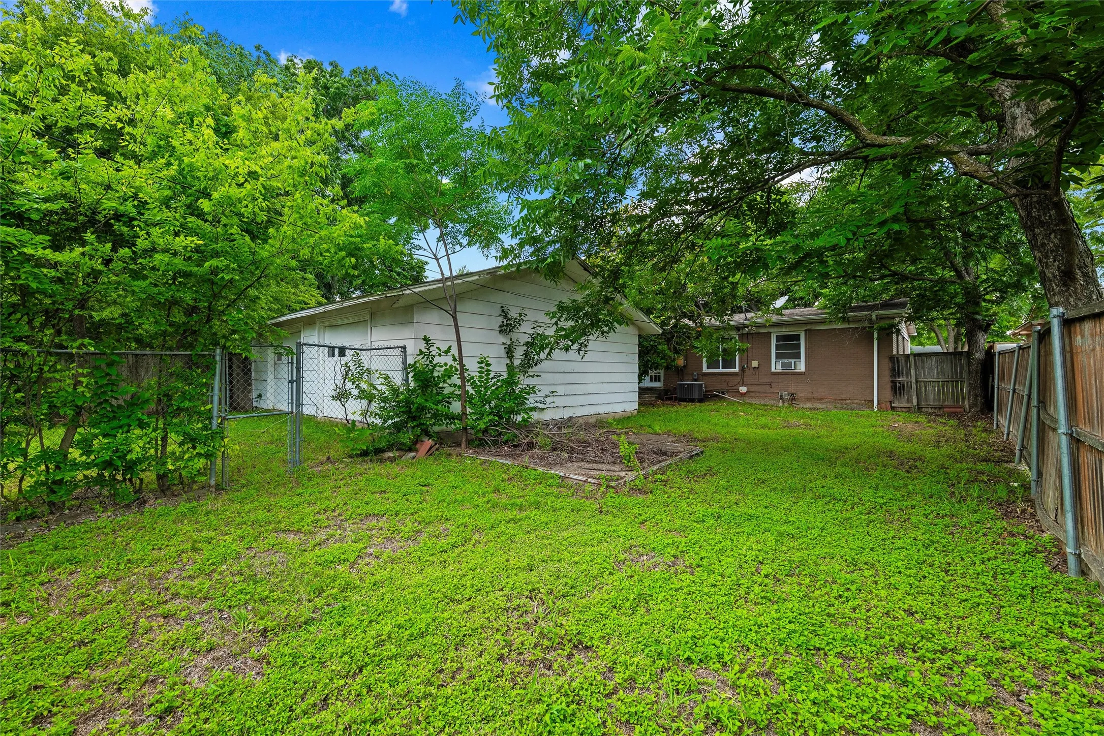 Backyard features detached storage building with exterior access. Brick exterior of the main residence is visible in the background. Wood and chain-link fencing enclose the yard. Grass lawn extends throughout.