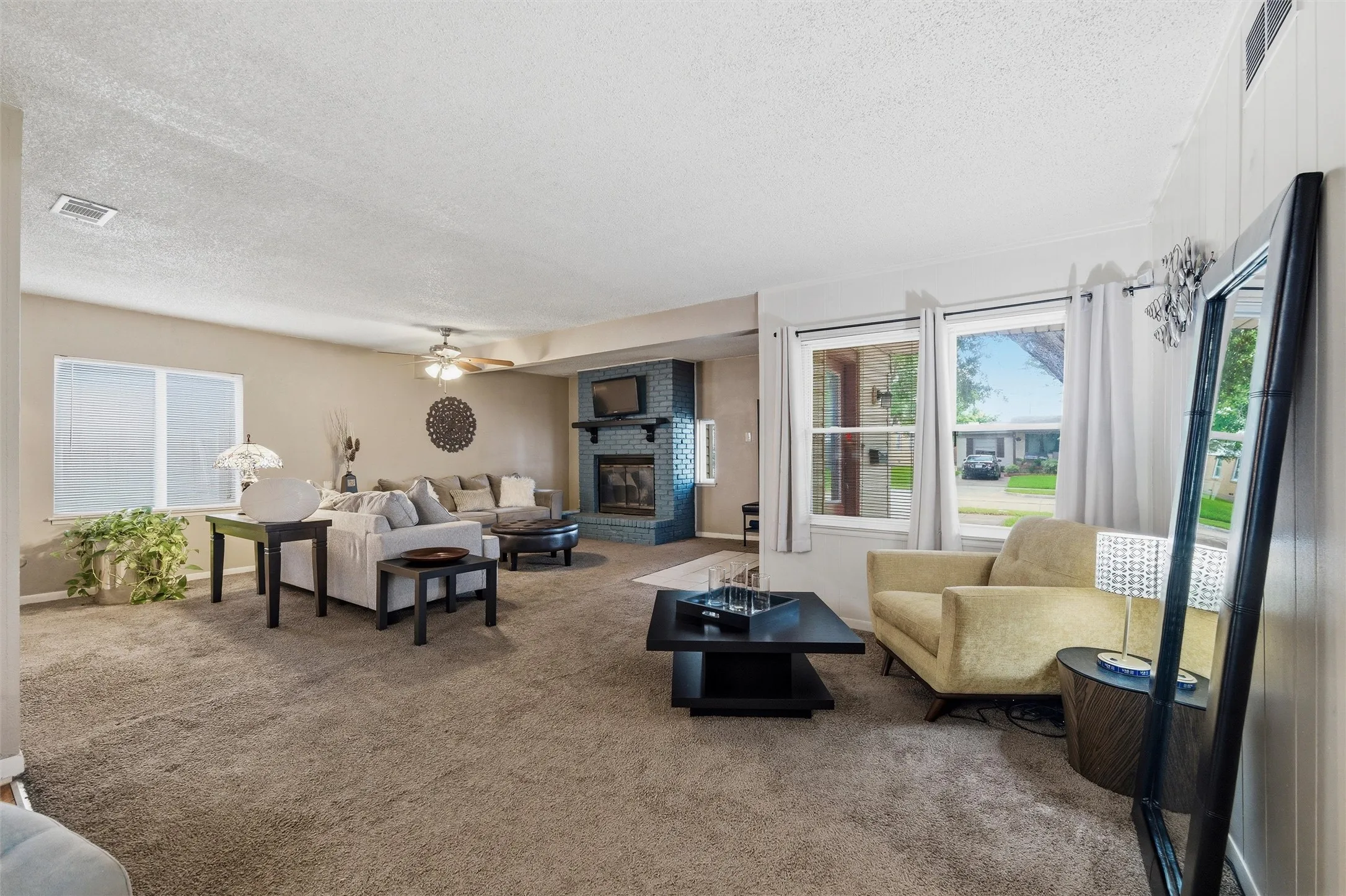 Living Room features a textured ceiling with ceiling fan and light fixture. Painted brick fireplace with raised hearth and wood mantel anchors the space. Windows provide natural light. Carpet flooring extends throughout.