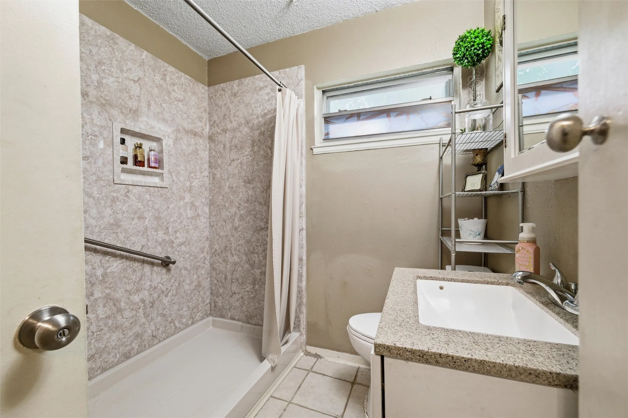 athroom features a textured ceiling with vanity lighting. Single-sink vanity includes laminate countertop, undermount sink, and medicine cabinet with mirror. Walk-in shower includes built-in shelving and grab bars. Window provides natural light. Tile flooring extends throughout.