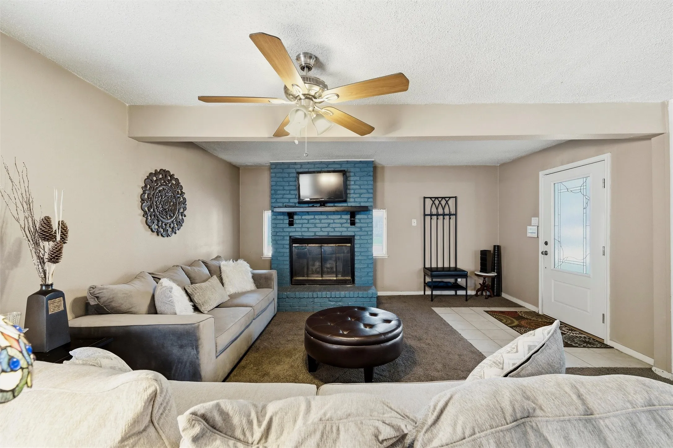 Living Room features a textured ceiling with ceiling fan and light fixture. Painted brick fireplace with raised hearth and wood mantel serves as the focal point of the room. Entry door includes decorative glass insert with sidelight detail. Carpet flooring extends throughout with tile flooring at the entry.