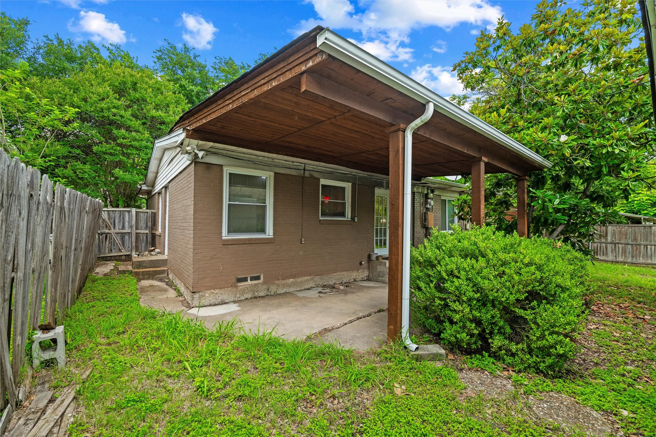 Rear Exterior / Carport features a wood-planked ceiling supported by wood posts. Brick exterior includes rear access door and multiple windows. Covered structure extends along the rear elevation. Concrete surface extends throughout.