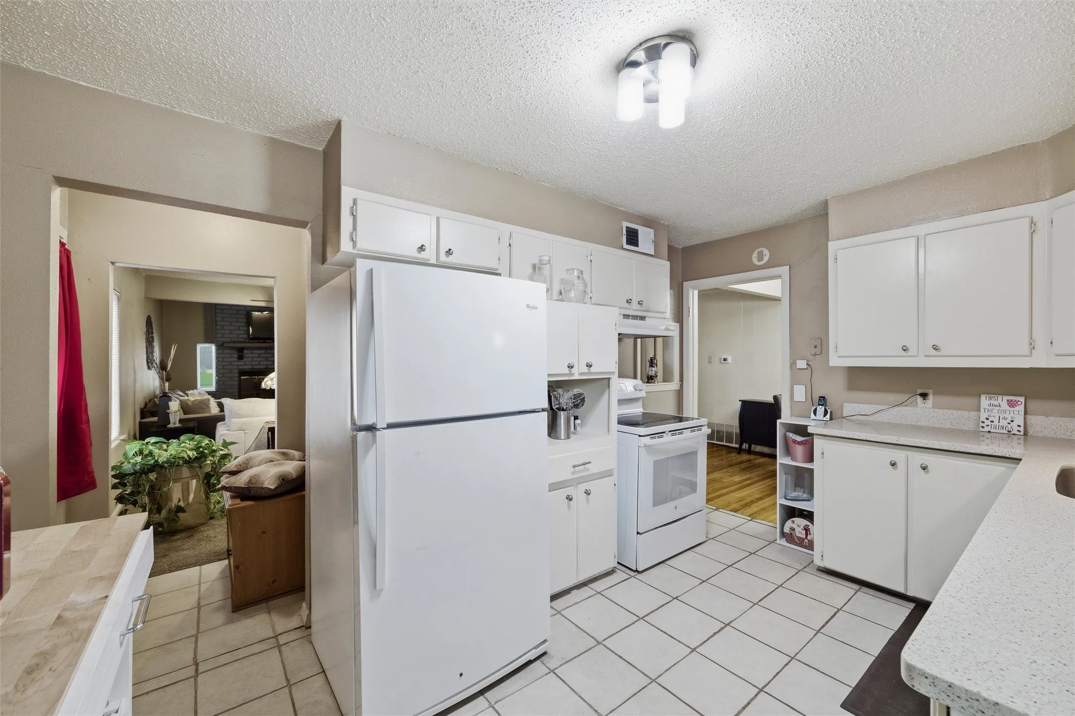 Kitchen features a textured ceiling with flush-mount light fixture. White raised-panel cabinetry extends throughout with laminate countertops. Appliances include an electric range and refrigerator. Open layout connects directly to the living room and dining area. Tile flooring extends throughout.