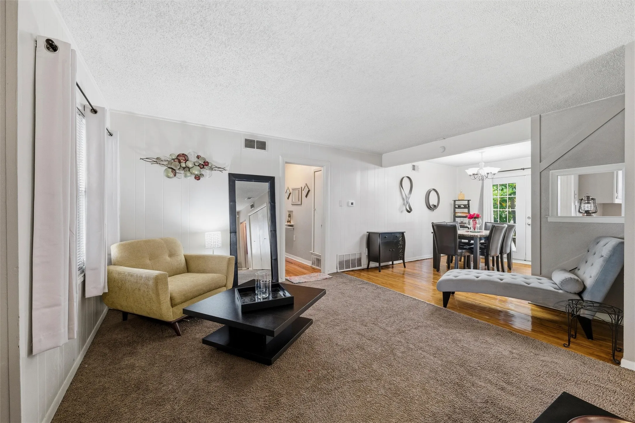 Living Room features a textured ceiling with recessed ventilation. Windows provide natural light throughout the room. Open layout connects directly to the dining area and hallway. Carpet flooring extends throughout.