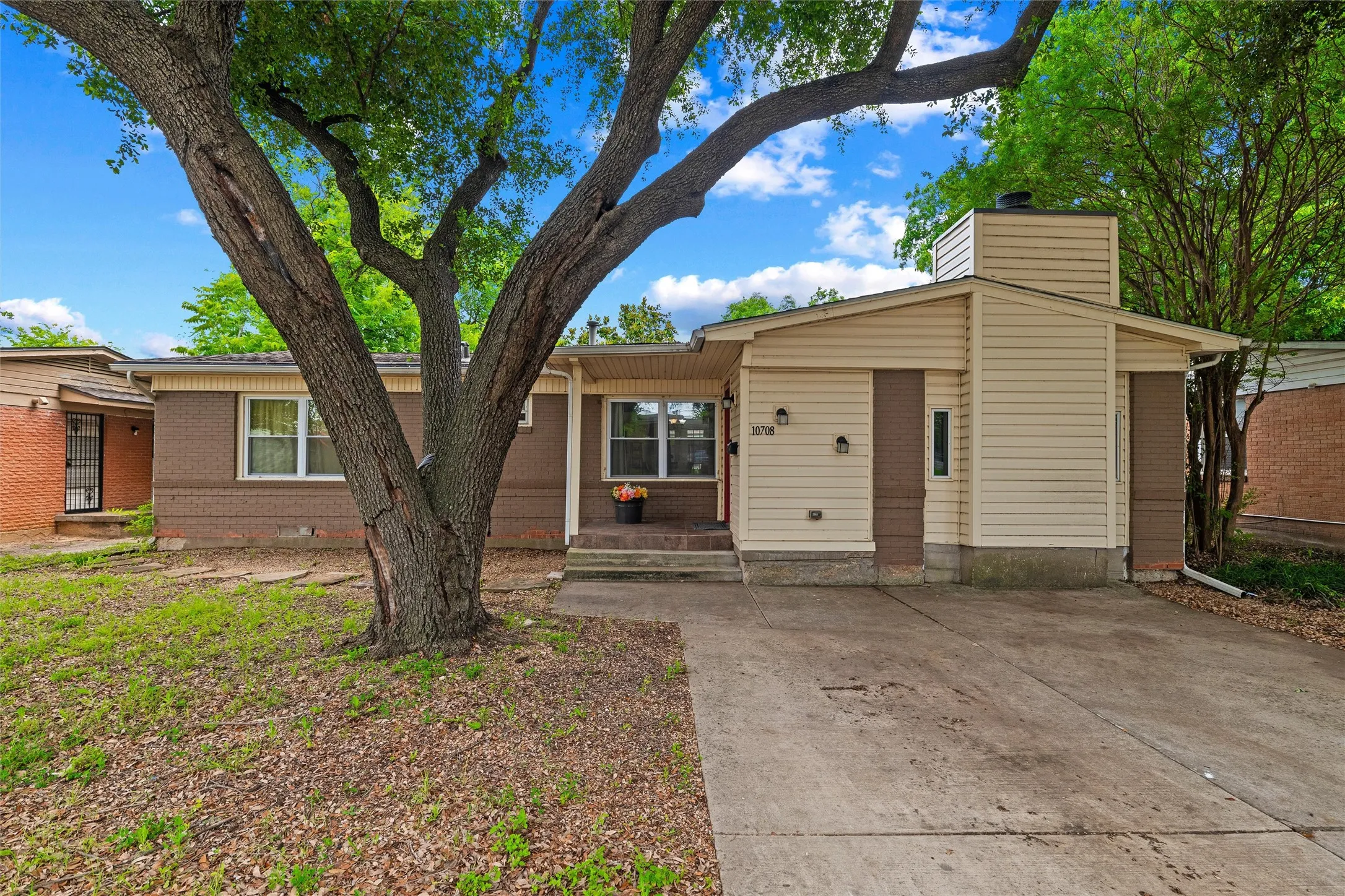Front Exterior features a composition roof with gutter system, painted brick construction, and vertical board siding. Covered entry includes stepped concrete access and wall-mounted exterior lighting. Concrete driveway provides off-street parking. Mature trees frame the front yard.