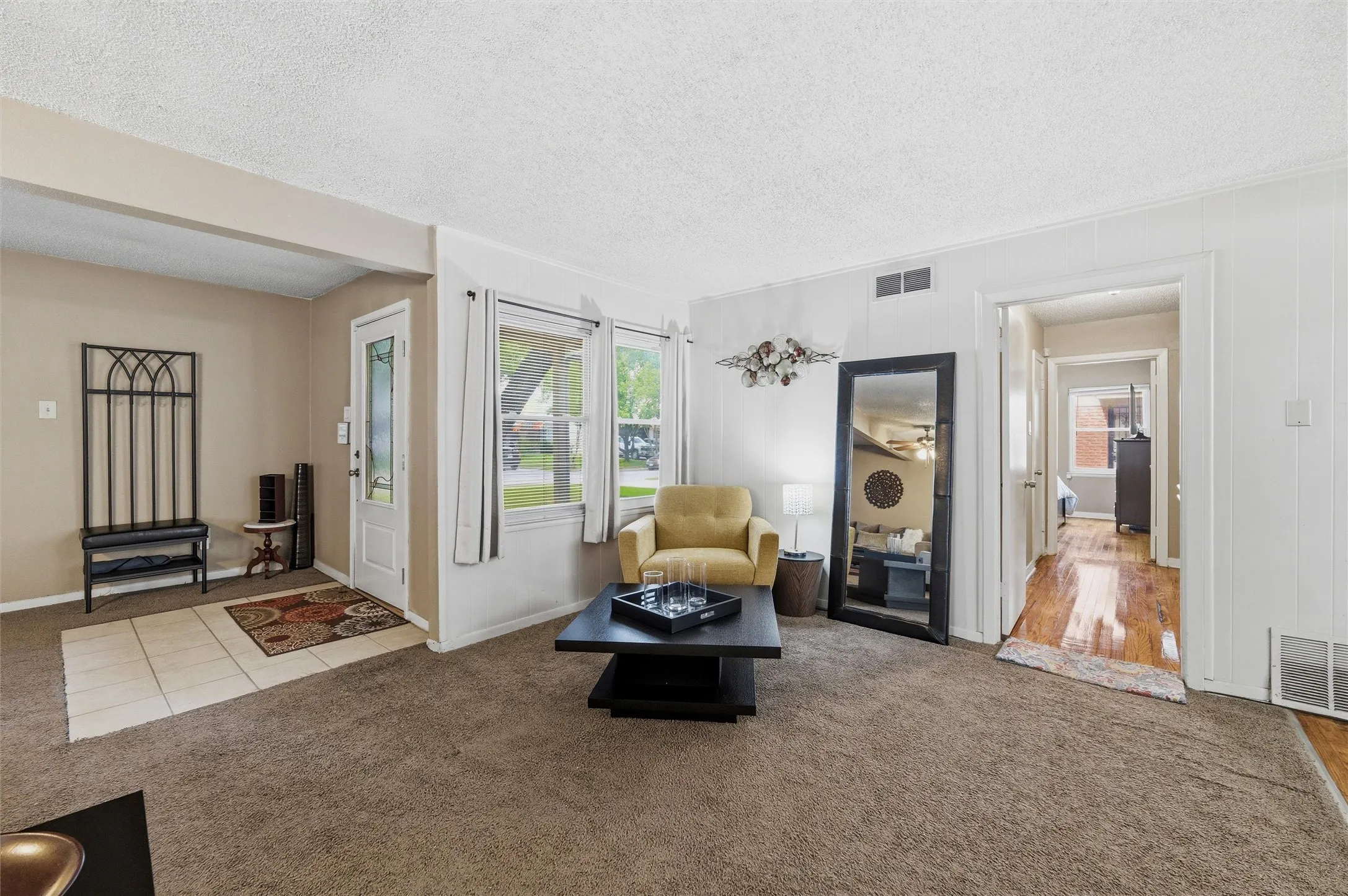 Living Room features a textured ceiling with recessed ventilation. Entry door includes decorative glass insert with sidelight detail. Front-facing windows provide natural light. Open layout connects directly to the hallway. Carpet flooring extends throughout with tile flooring at the entry.