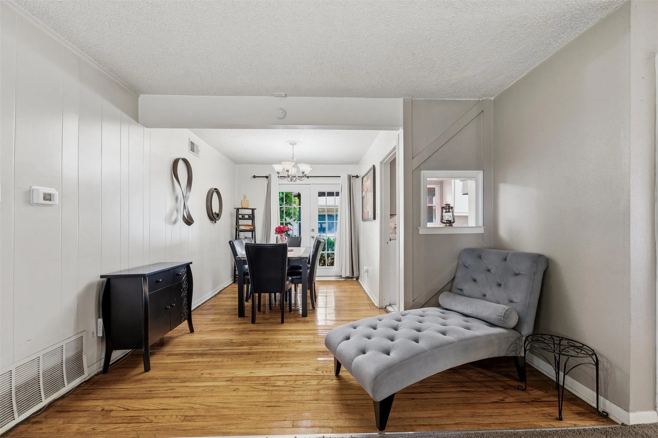 Dining Area features a textured ceiling with chandelier light fixture. Vertical board wall paneling accents the wall space. Pass-through opening connects directly to the kitchen. French doors provide natural light and exterior access. Hardwood flooring extends throughout.
