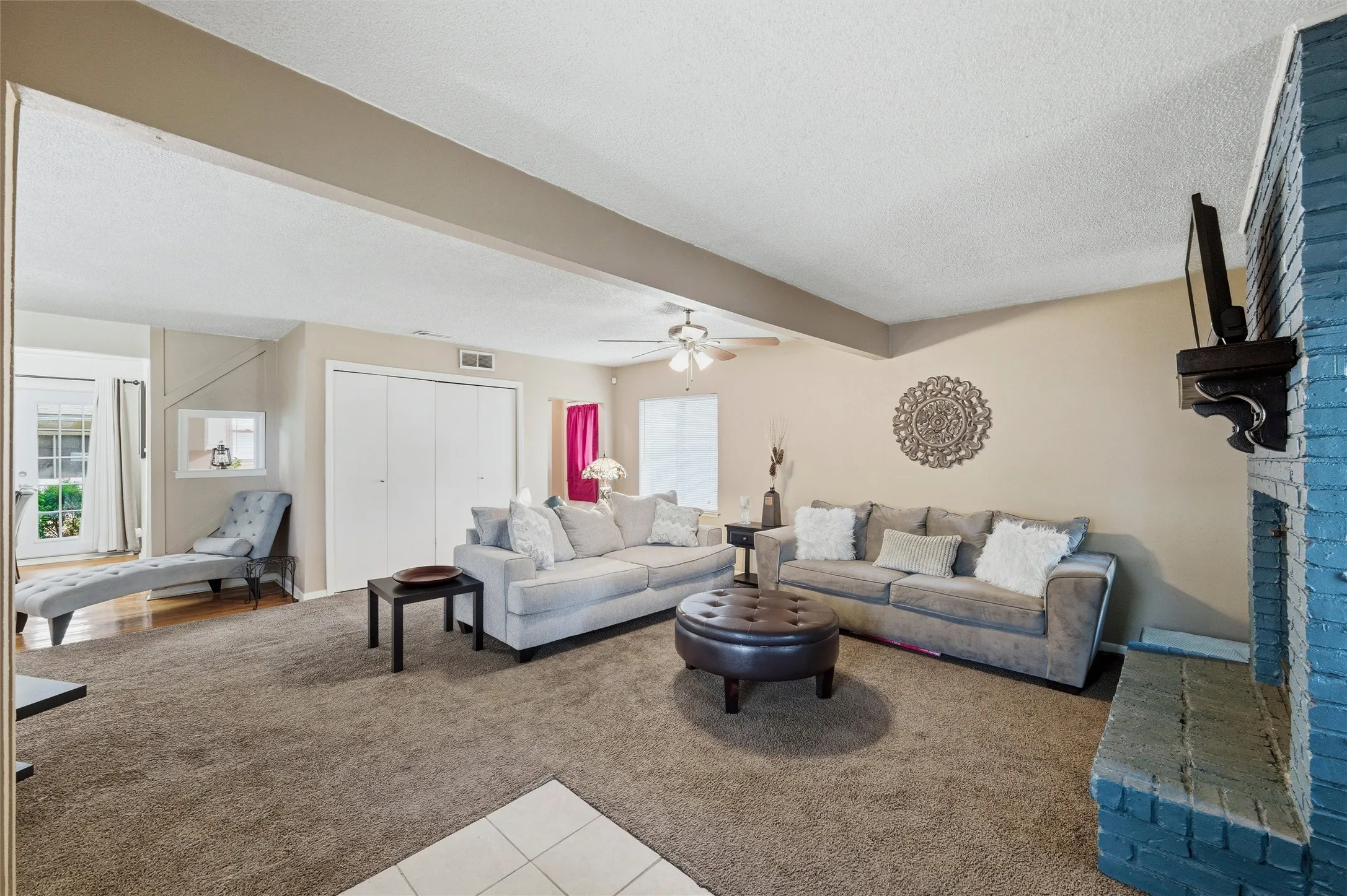 Living Room features a textured ceiling with ceiling fan and light fixture. Painted brick fireplace with raised hearth and wood mantel anchors the space. Windows provide natural light. Open layout connects directly to the dining area. Carpet flooring extends throughout.