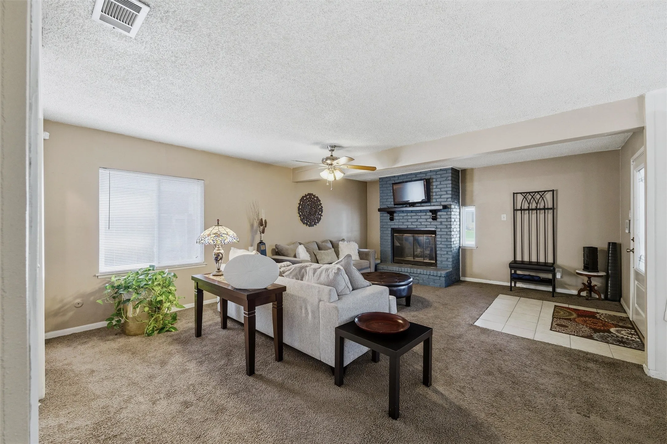 Living Room features a textured ceiling with ceiling fan and light fixture. Entry door includes decorative glass insert. Painted brick fireplace with raised hearth and wood mantel remains visible from this angle. Carpet flooring extends throughout with tile flooring at the entry.